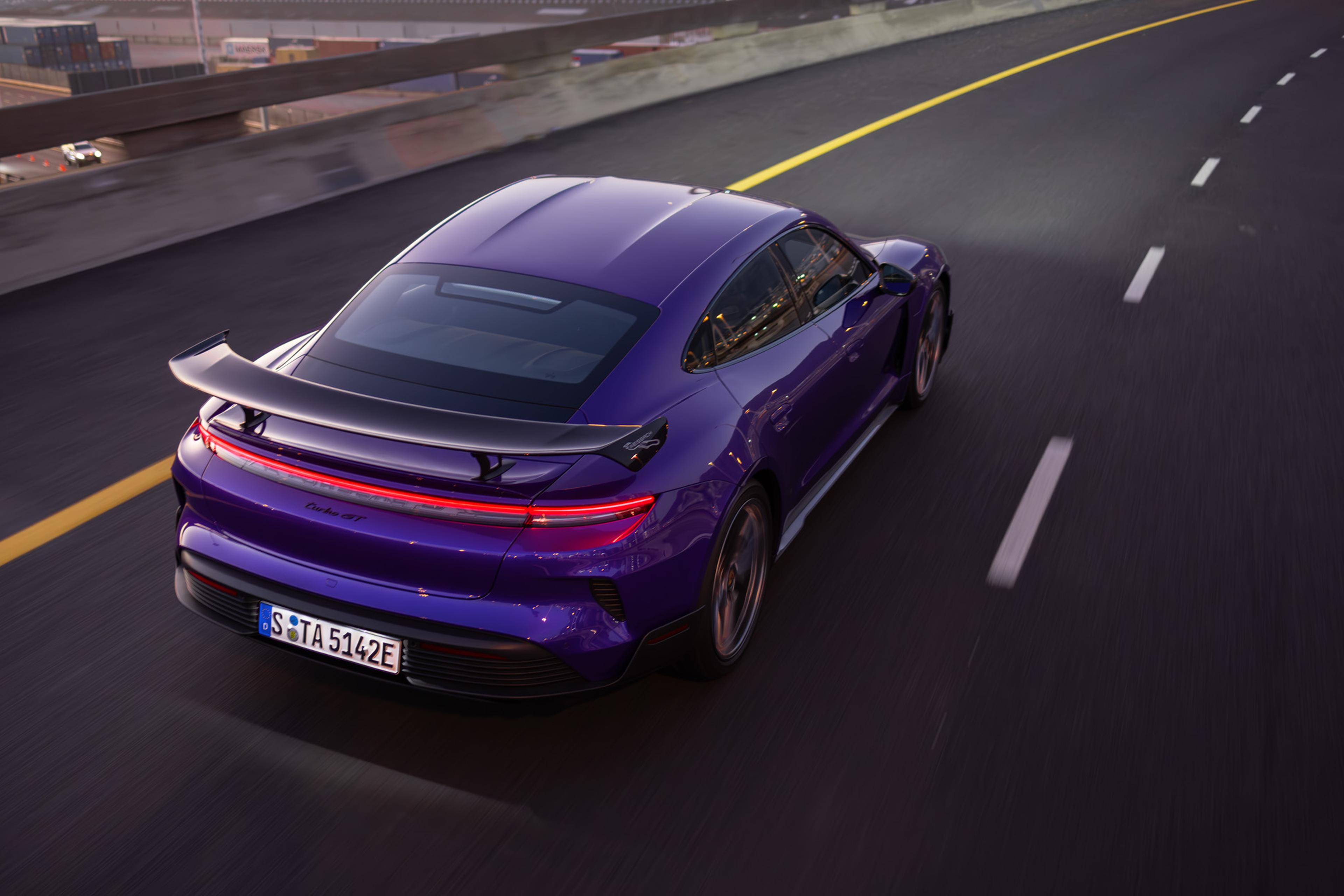 A purple porsche taycan with a spoiler drives on a highway, surrounded by a cityscape, during dusk.