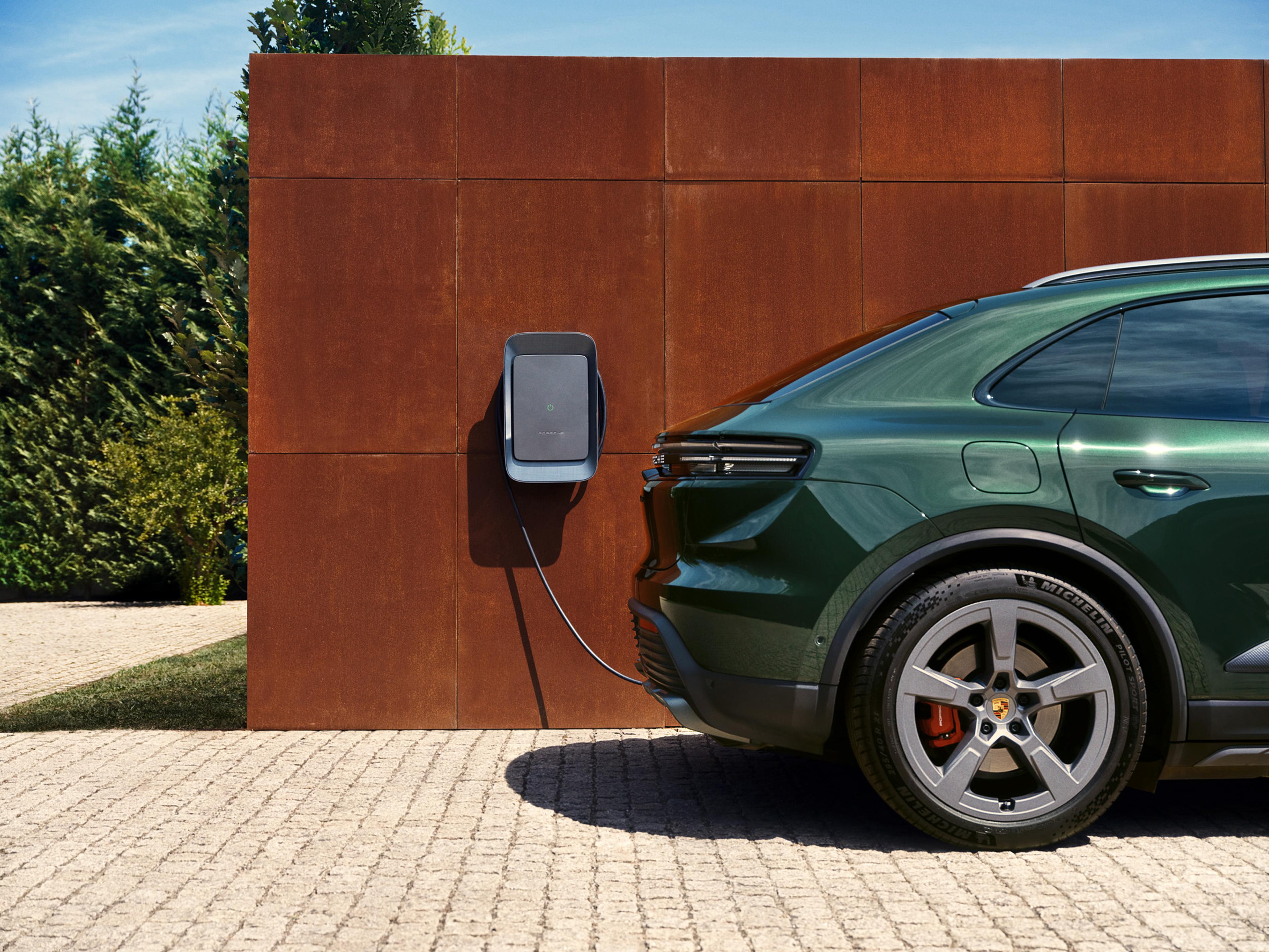 Ein grünes Elektroauto lädt an einer Wandladestation neben einer rostigen Metallplatte; im Hintergrund sind Grünflächen und ein klarer Himmel zu sehen.