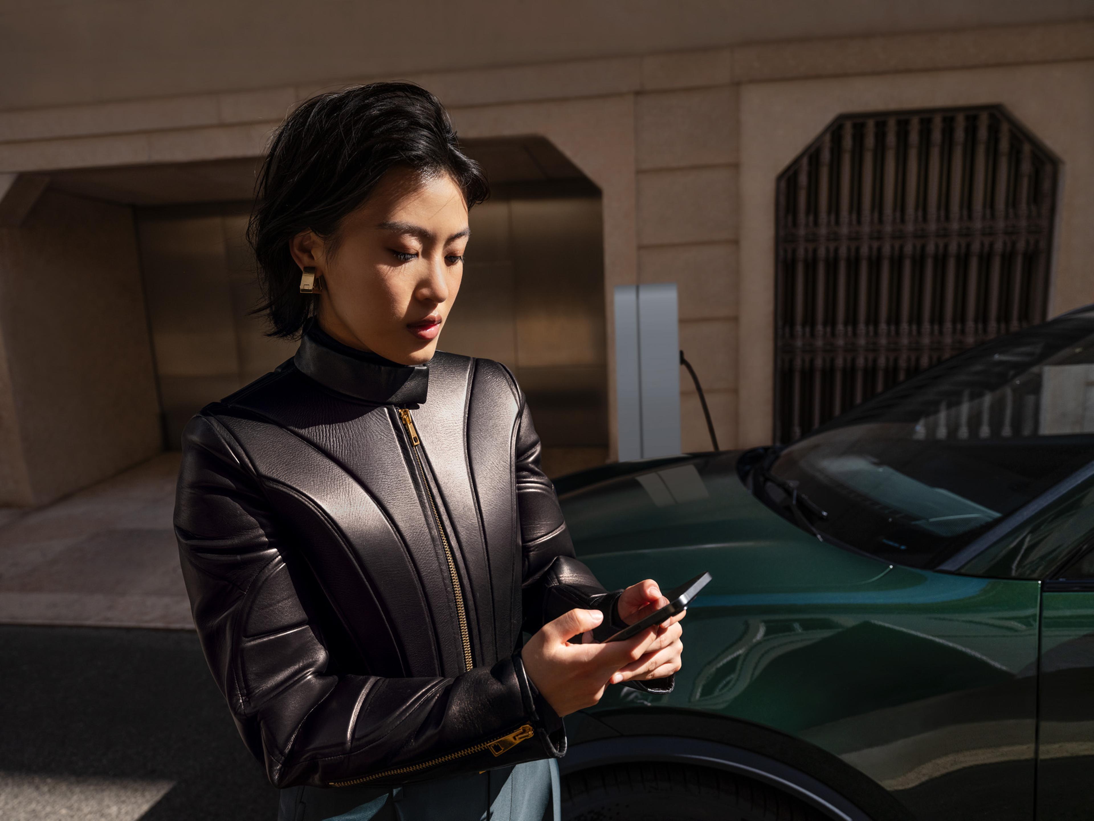 Eine Person in einer schwarzen Lederjacke benutzt ein Smartphone neben einem grünen Auto in einer städtischen Umgebung.