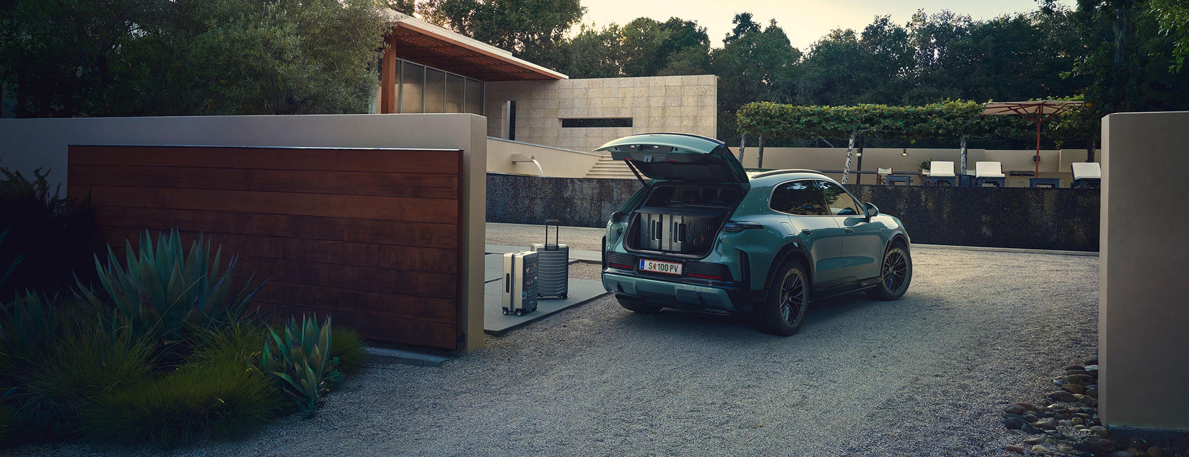 Ein blaugrüner Porsche Cayenne mit geöffnetem Kofferraum steht auf einer modernen Auffahrt, flankiert von zwei Gepäckstücken, mit einem modernen Haus im Hintergrund.