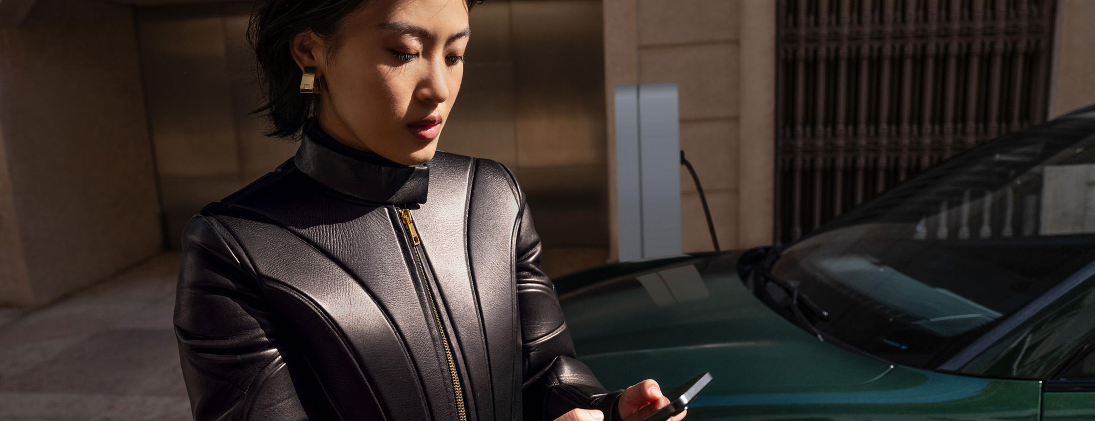 Eine Frau in einer schlichten schwarzen Lederjacke blickt auf ihr Handy; sie steht in der Nähe eines grünen Autos, im Hintergrund ist eine Ladestation zu sehen.