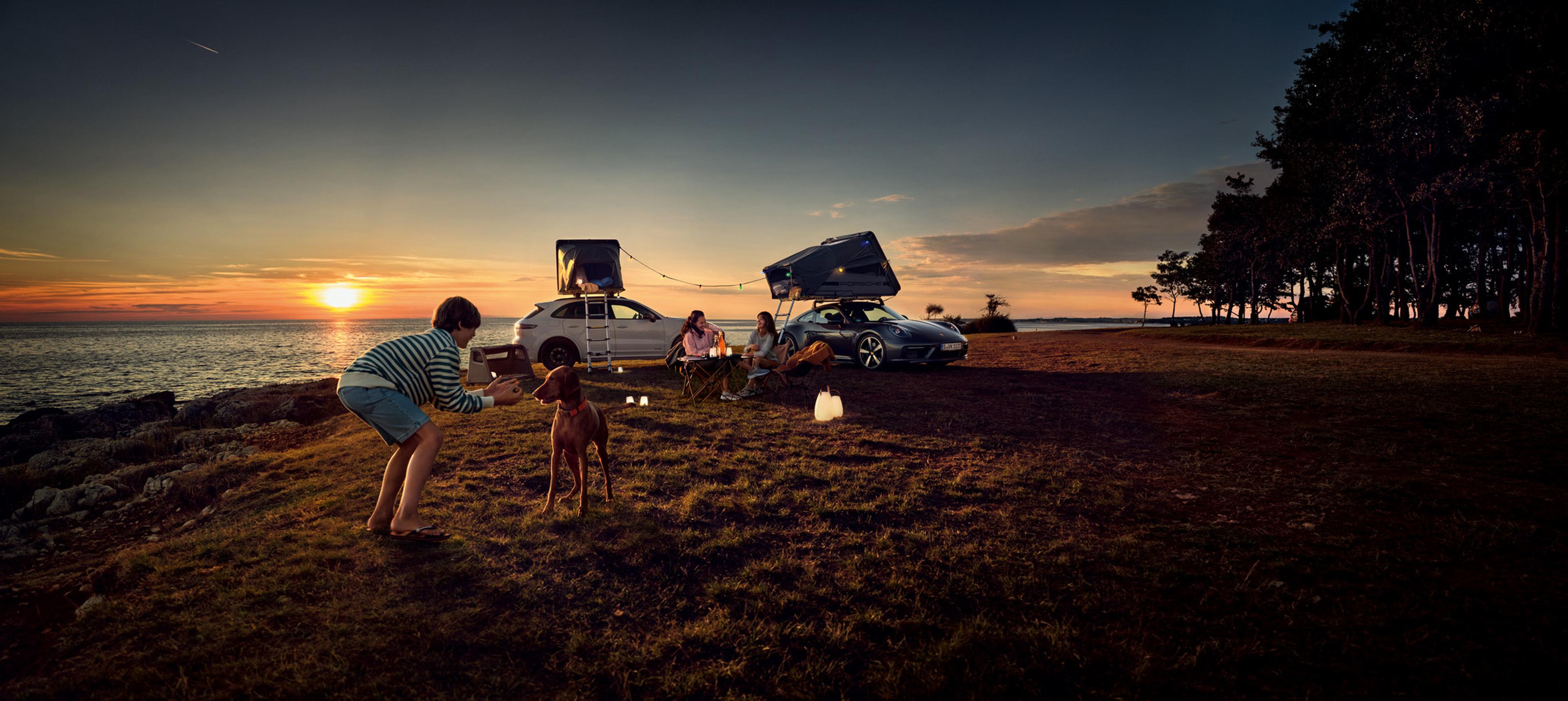 Family by the ocean at sunset with a Porsche Cayenne GTS and Porsche 911 Carrera 4 parked nearby.