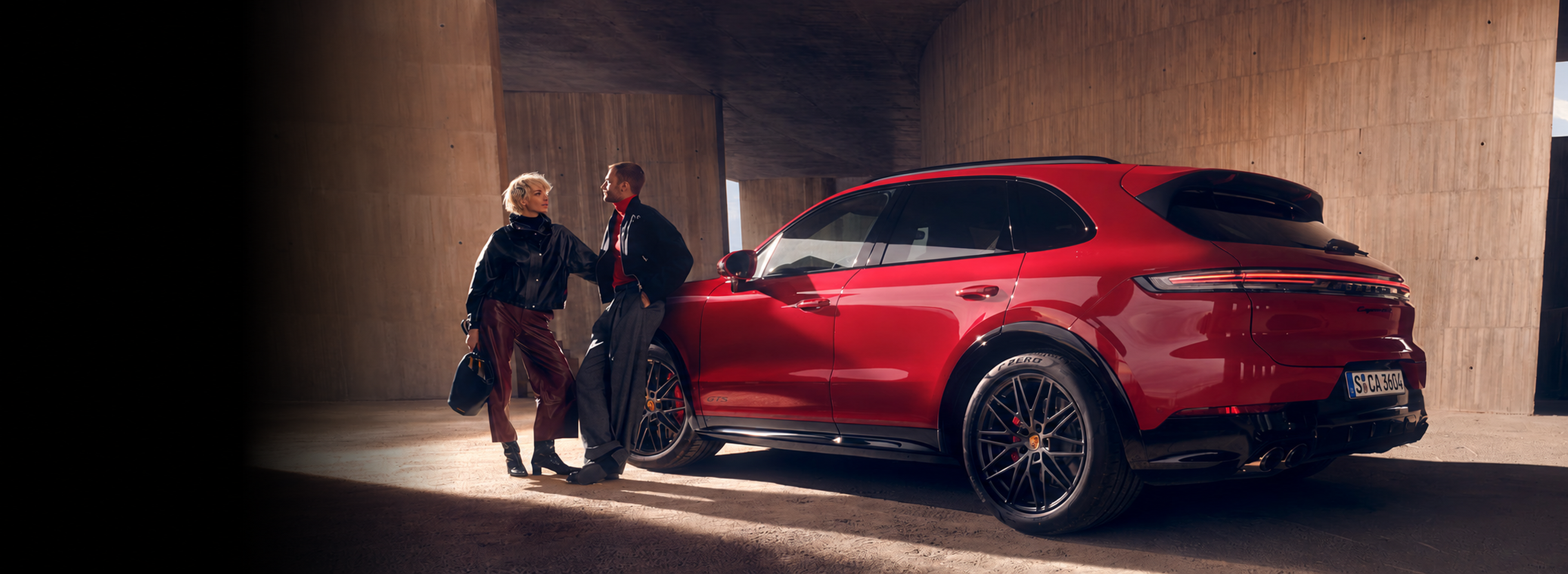 A man and woman stand beside a red luxury SUV in a concrete setting, engaging in conversation.