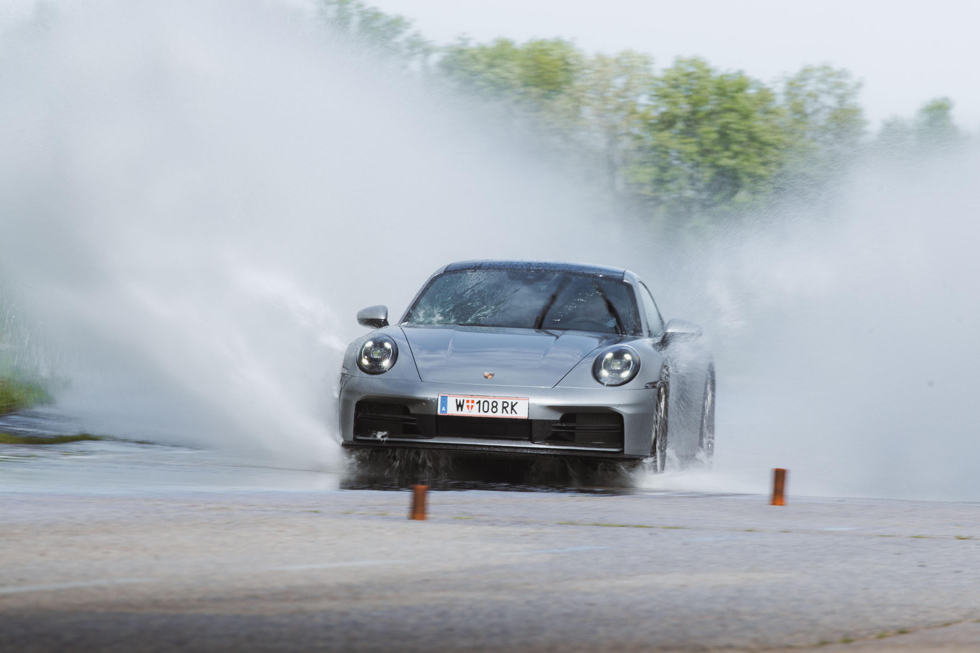 Ein silberner Sportwagen fährt durch Wasser auf einer nassen Straße und wirbelt dabei zu beiden Seiten große Wassermengen auf; im Hintergrund sind Bäume zu sehen.