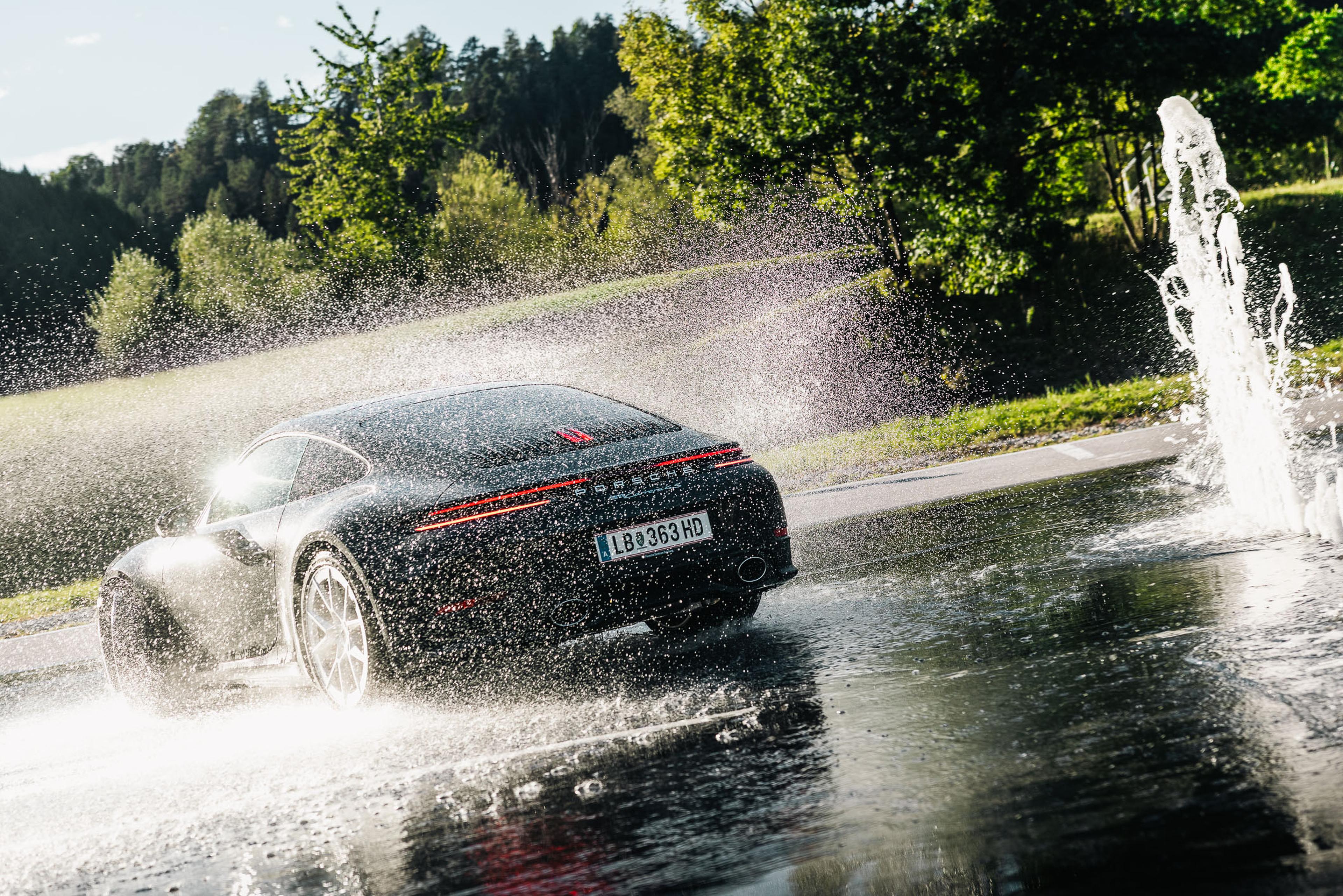 Ein eleganter schwarzer Sportwagen fährt auf nasser Straße durch aufspritzendes Wasser; das Sonnenlicht schimmert, umgeben von grünen Bäumen.