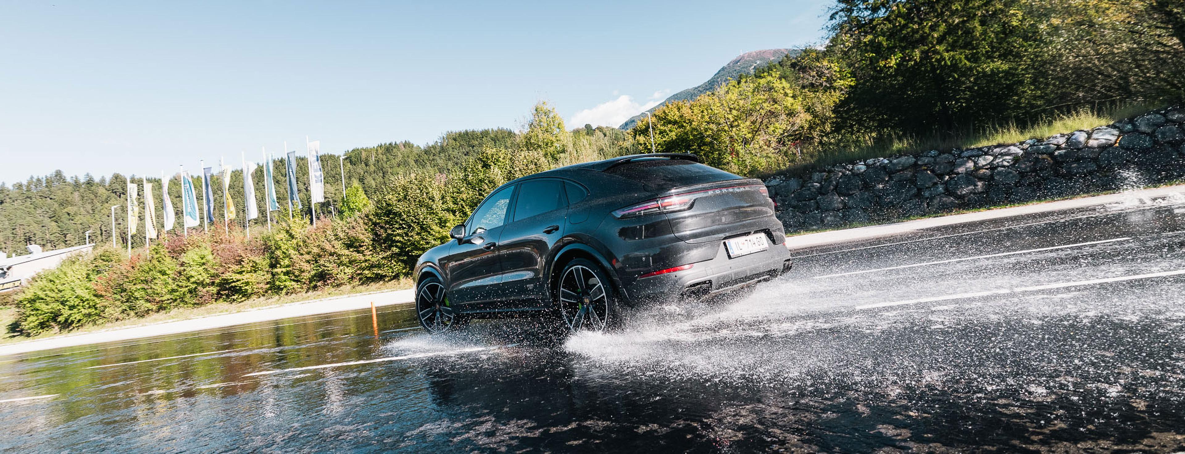 Ein schwarzer SUV fährt auf einer nassen Straße, wobei Wasser aufspritzt; im Hintergrund sind Bäume und Flaggen zu sehen.