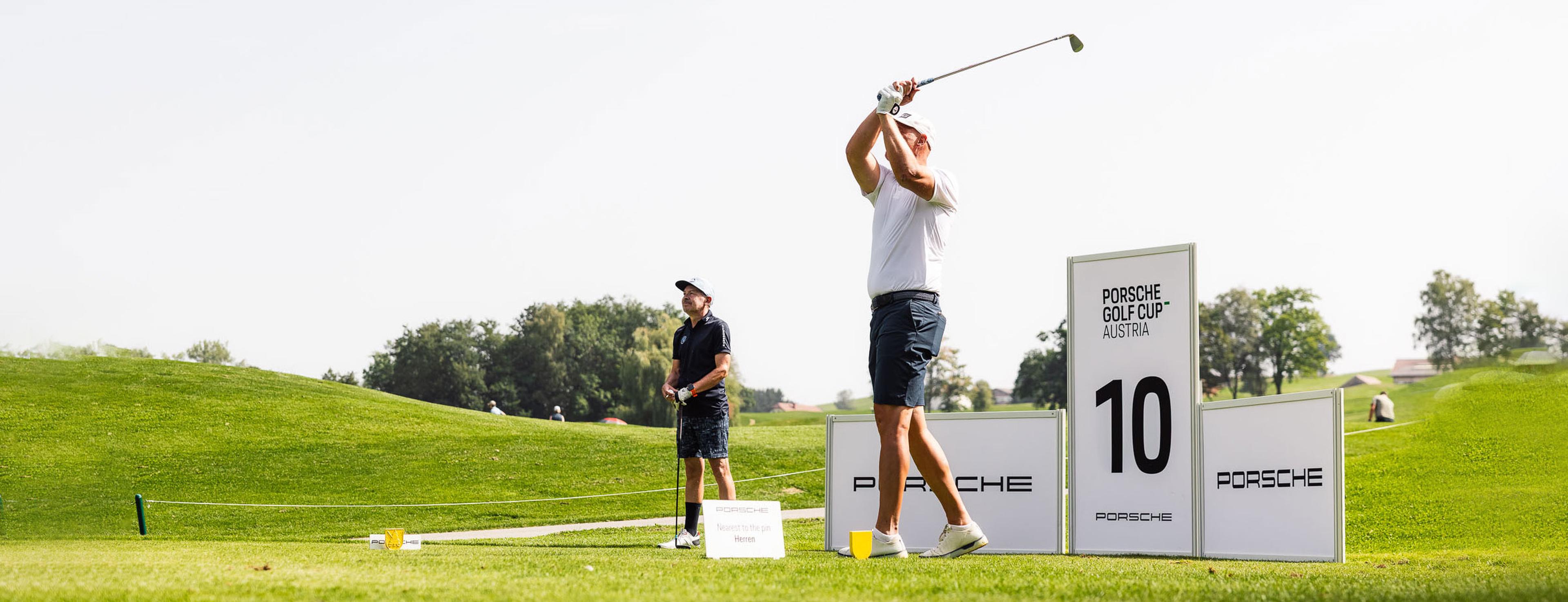 Zwei Männer auf einem Golfplatz während eines Turniers: Einer schwingt den Schläger, der andere schaut zu. Schilder zeigen die Aufschrift „Porsche Golf Cup Austria“. Üppig grüne Landschaft.