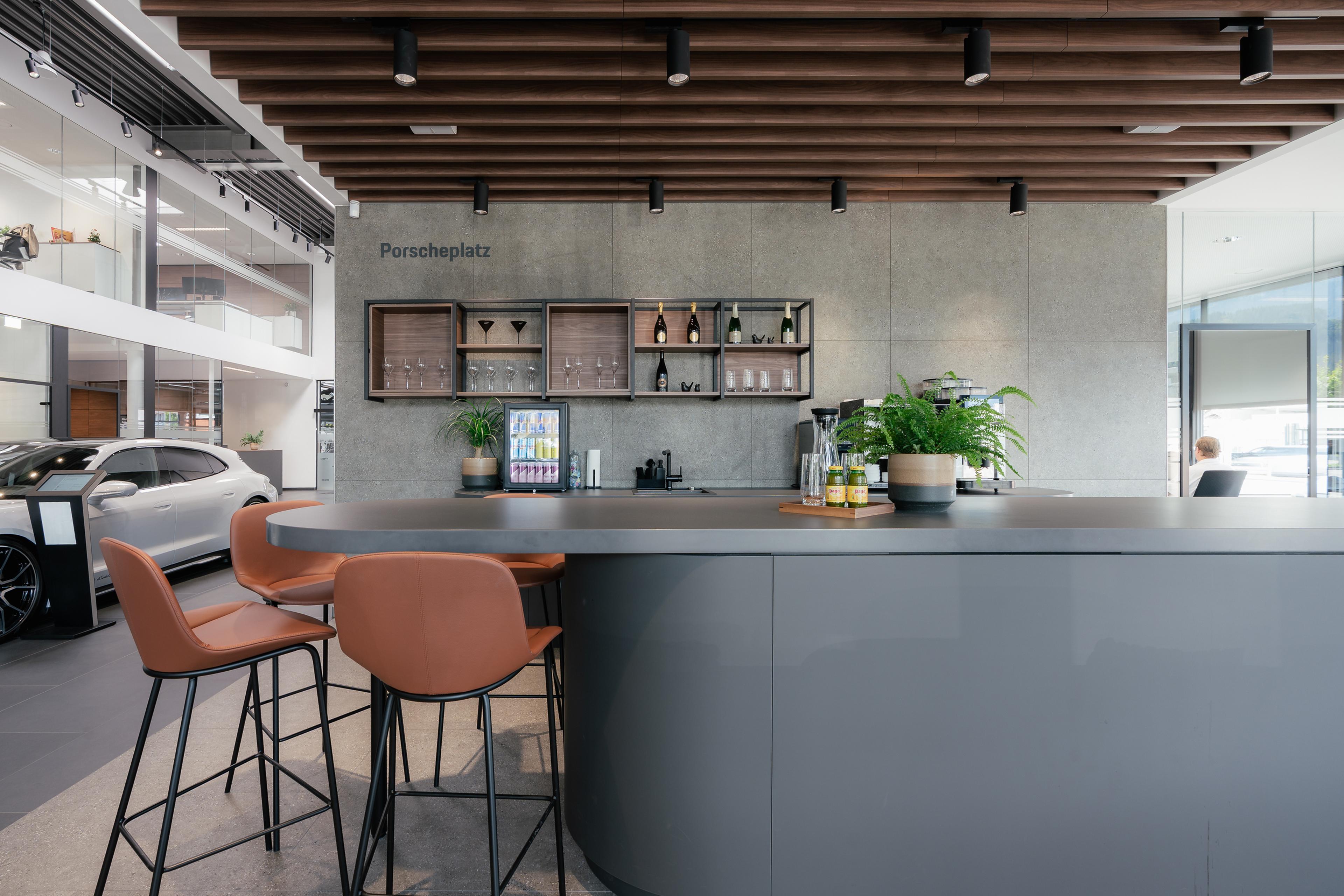 Modern showroom cafe area with sleek counter, orange barstools, display shelves, potted plants, and a silver sports car visible in the background.