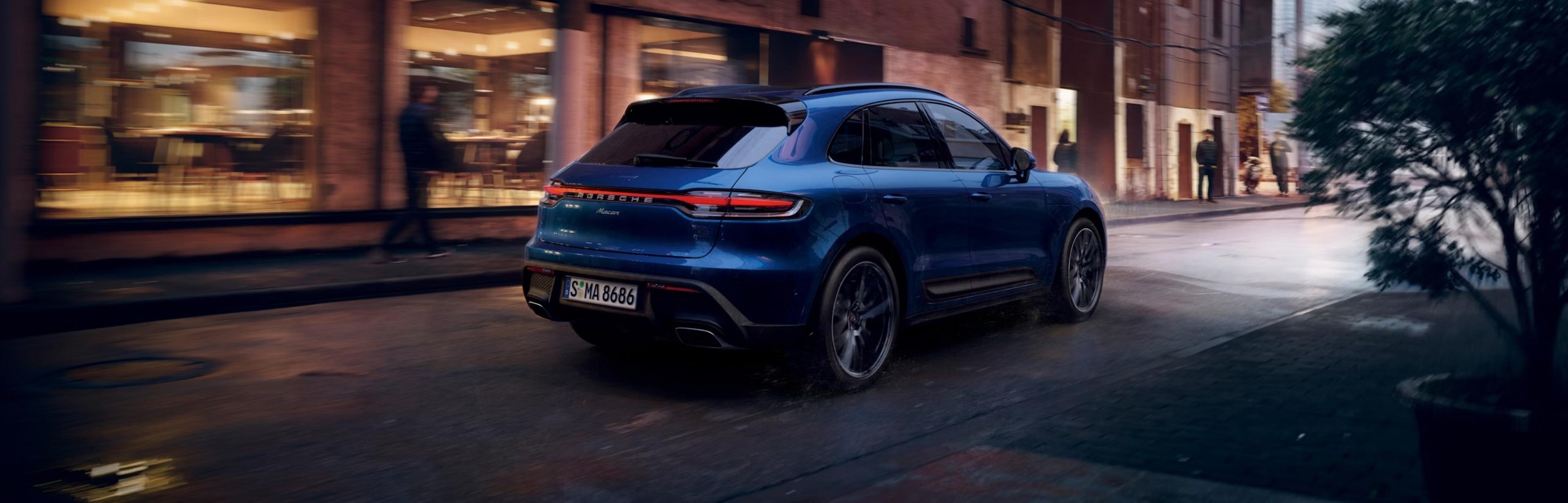 A blue SUV driving through a wet city street at night, with illuminated shop windows and blurred pedestrians in the background.