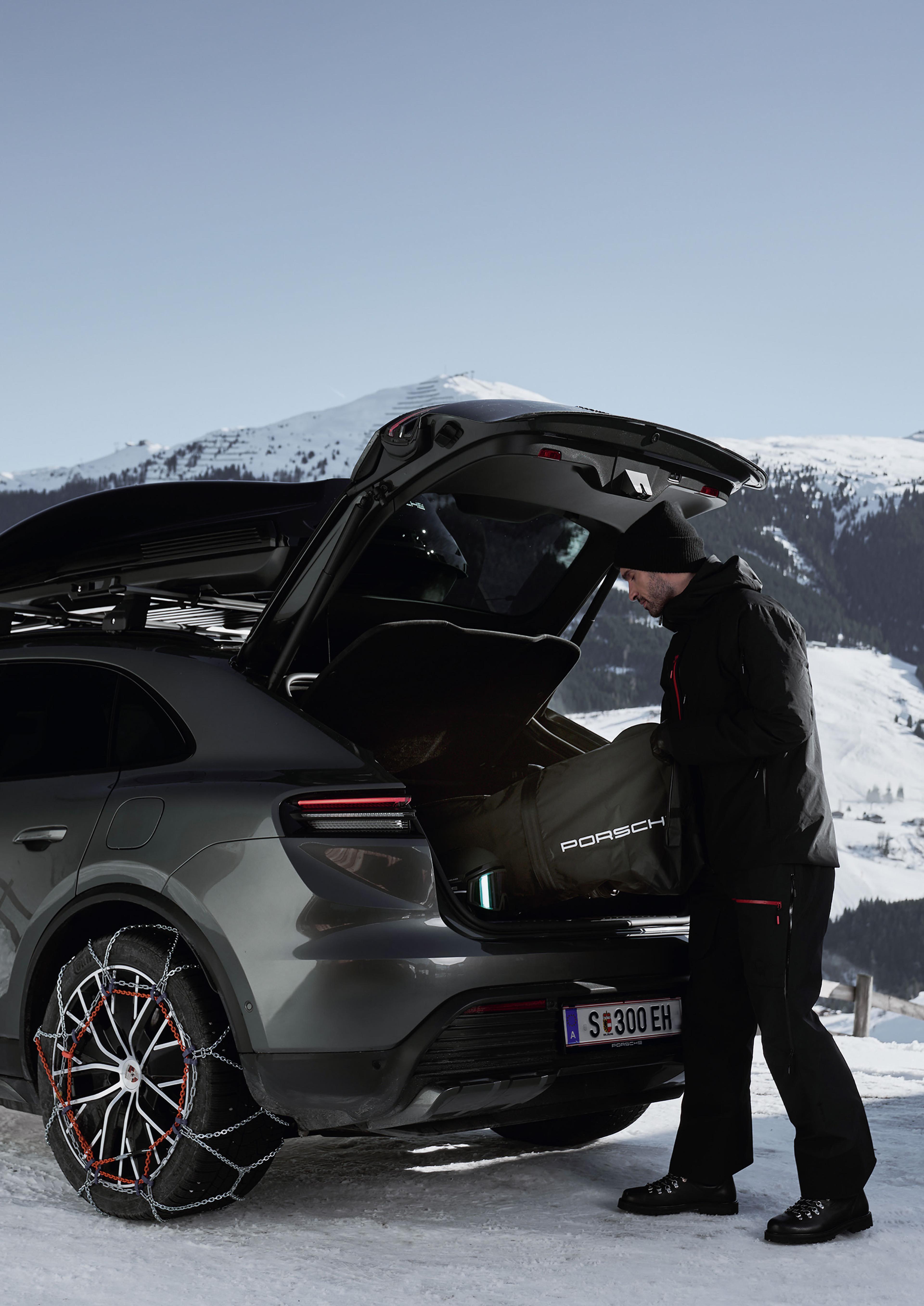 Eine in Winterkleidung gekleidete Person lädt eine Porsche-Tasche in den Kofferraum eines Autos, das auf verschneitem Untergrund vor einer Bergkulisse geparkt ist. Die Reifen sind mit Schneeketten ausgestattet.
