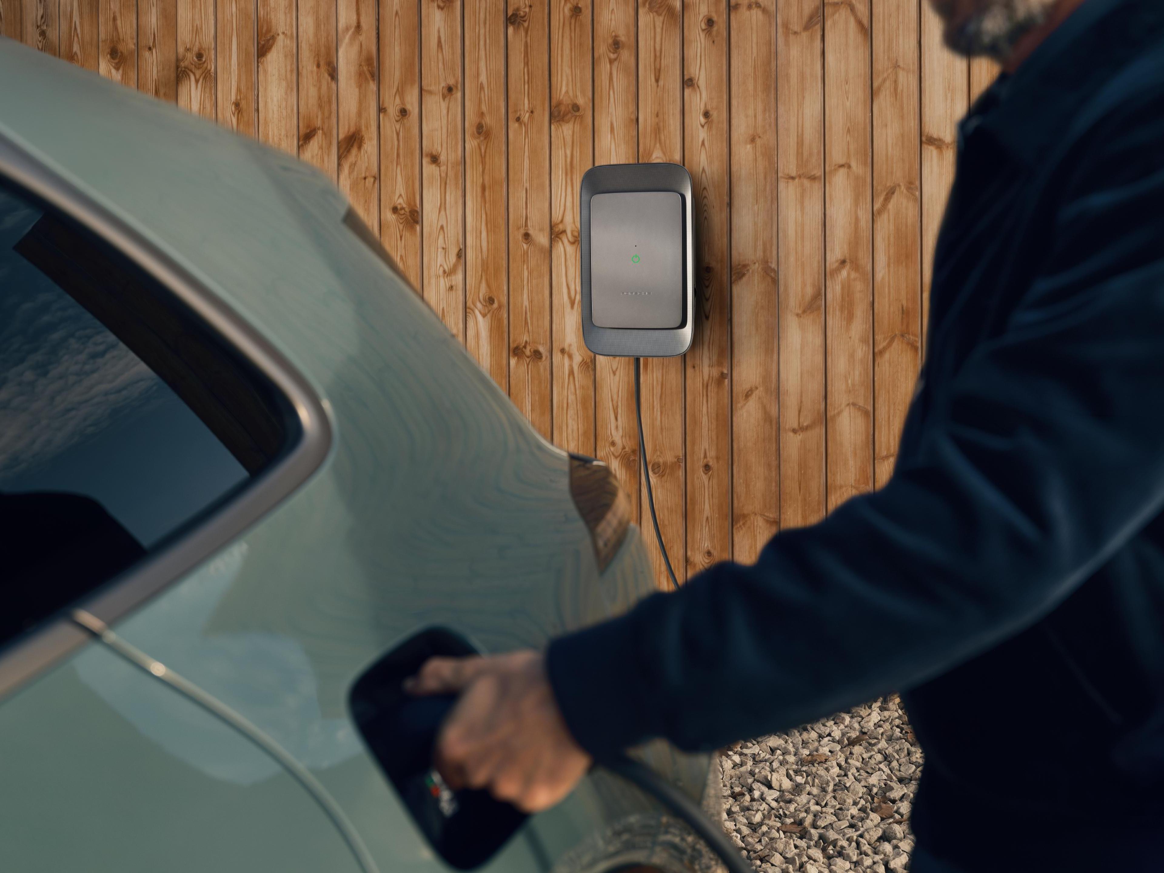 Eine Person schließt ein Elektroauto an eine an einer Holzwand montierte Ladestation an; der Untergrund besteht aus Kies.