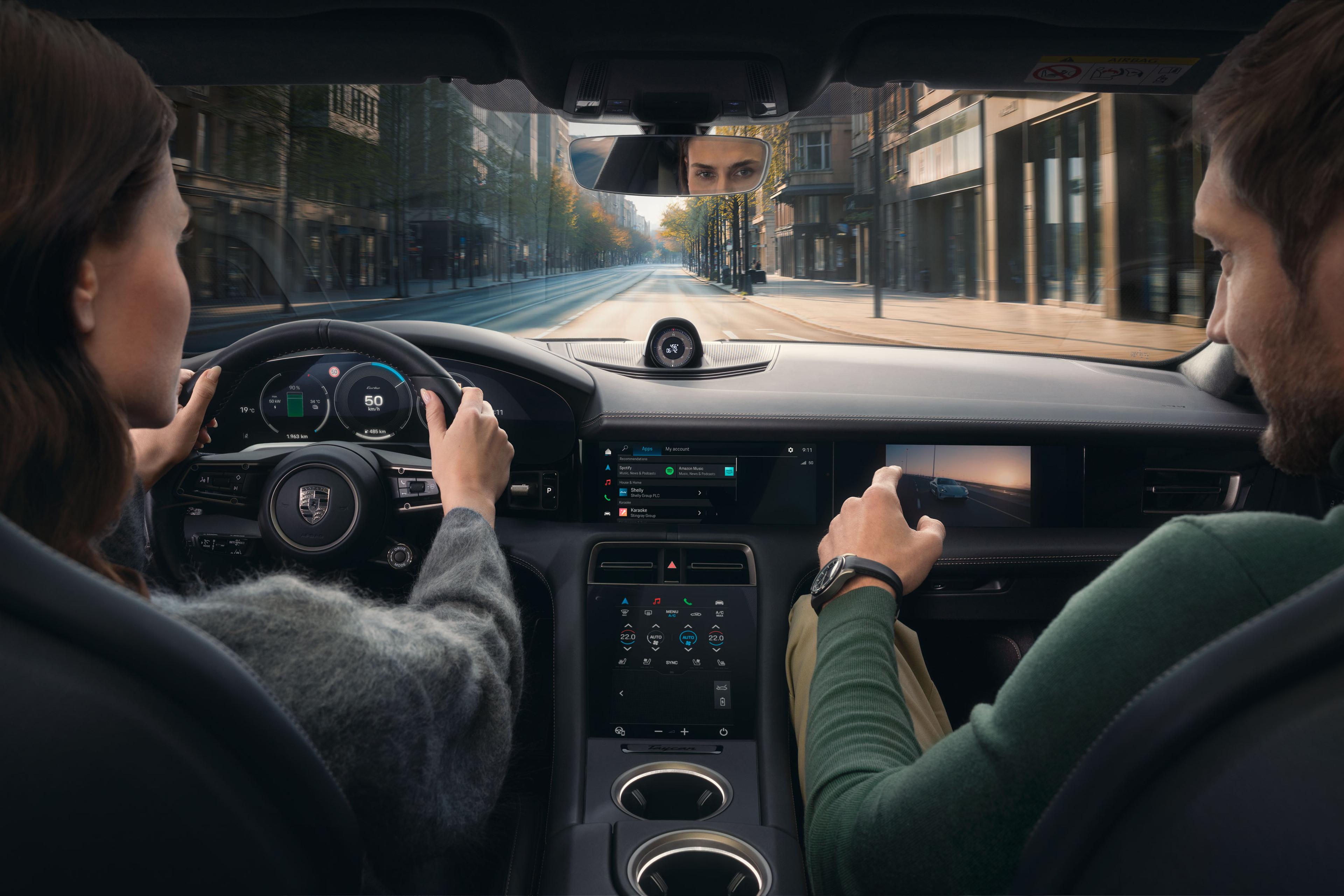 Fahrer und Beifahrer in einem Auto, der Fokus liegt auf den Anzeigen des Armaturenbretts; die Straße vor ihnen ist leer und von Gebäuden gesäumt.