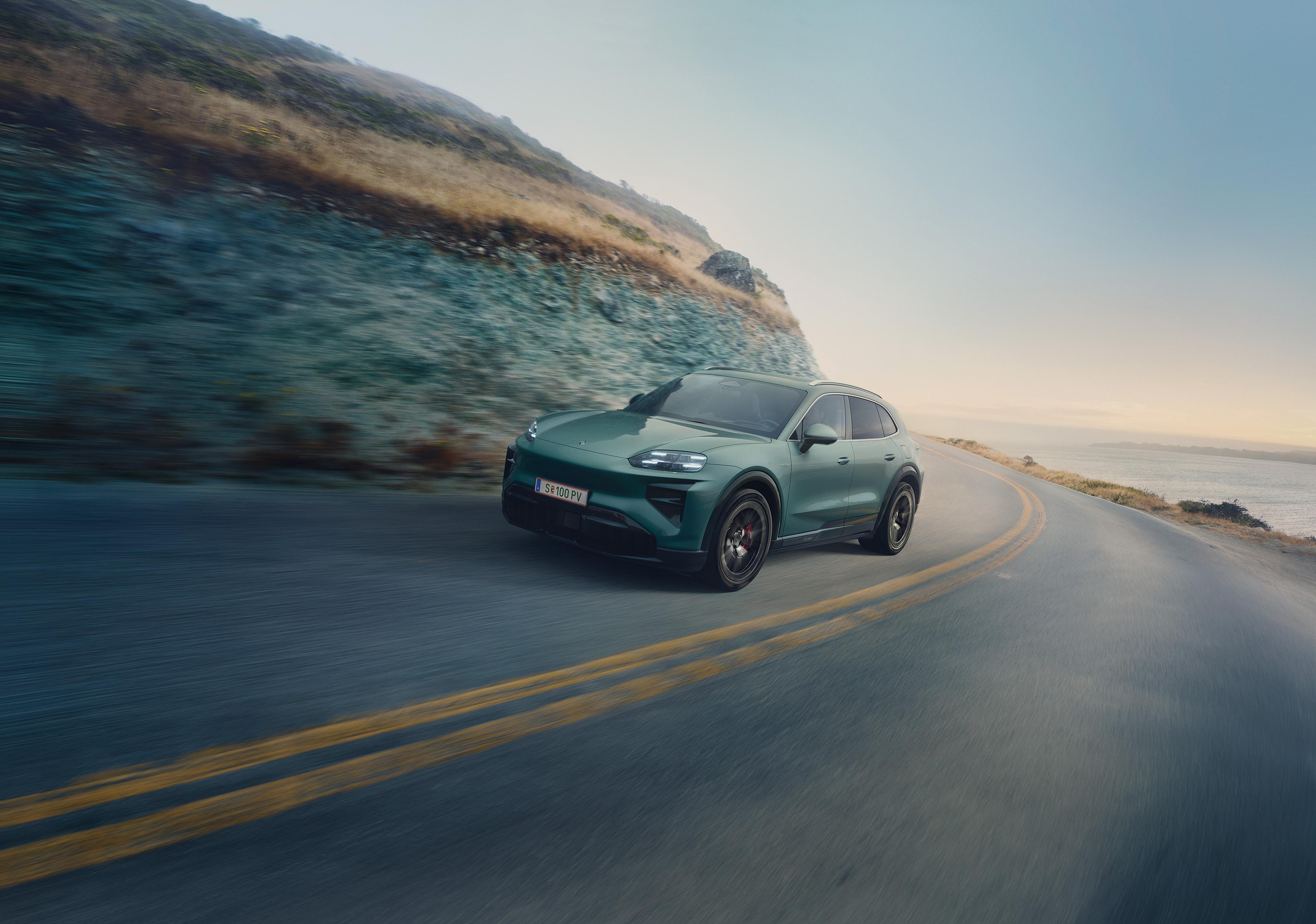 Ein grüner Porsche Cayenne mit Elektroantrieb fährt auf einer kurvenreichen Küstenstraße; im Hintergrund sind Klippen und das Meer unter klarem Himmel zu sehen.