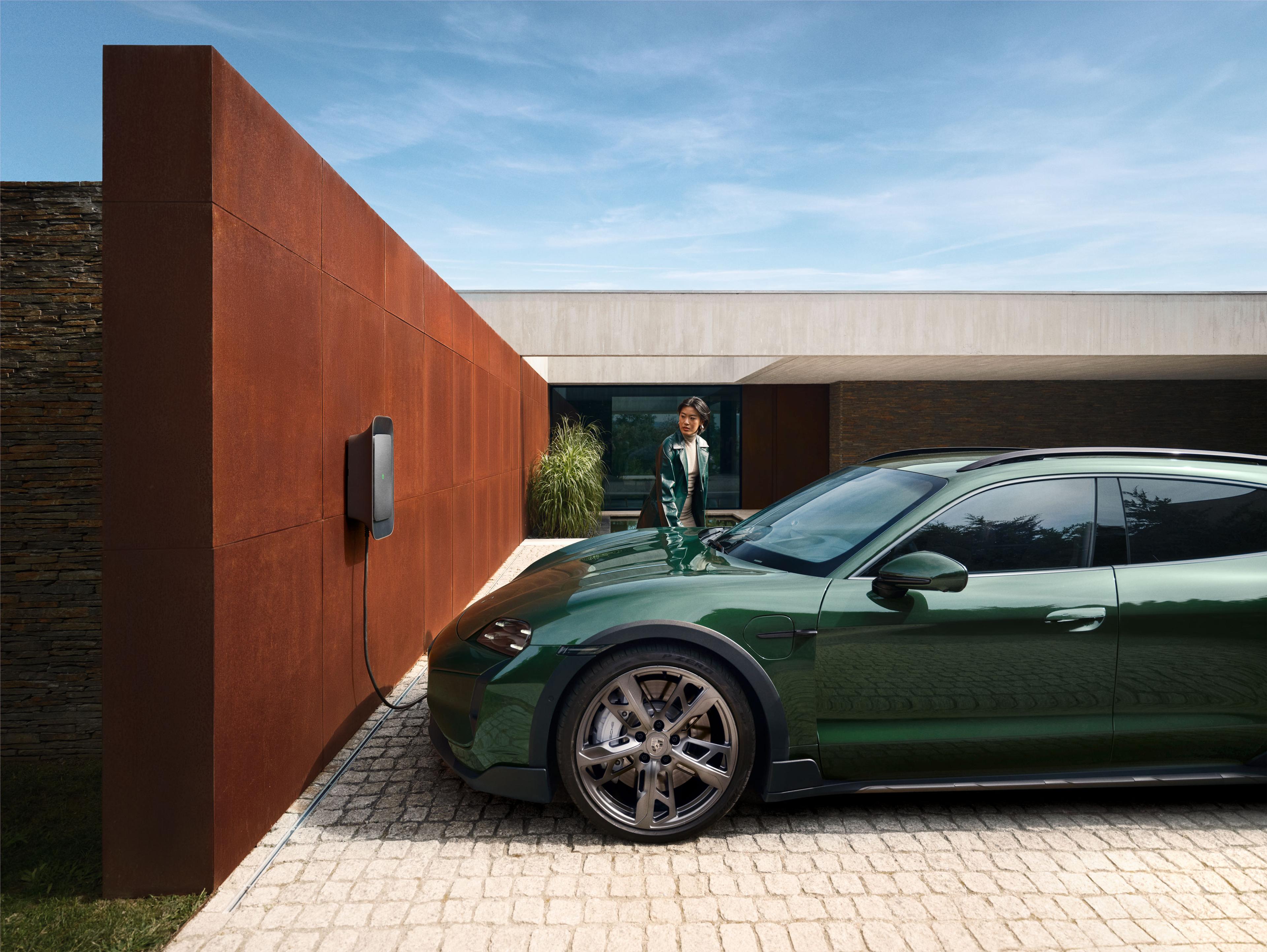 Ein grünes Elektroauto parkt und lädt vor einem Haus mit einer modernen Backsteinmauer unter einem klaren blauen Himmel. Eine Person steht in der Nähe.