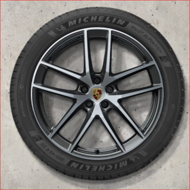 A black Macan rim with a Michelin tire with a dark gray multi-spoke alloy wheel, featuring a Porsche logo at the center, on a light concrete surface.