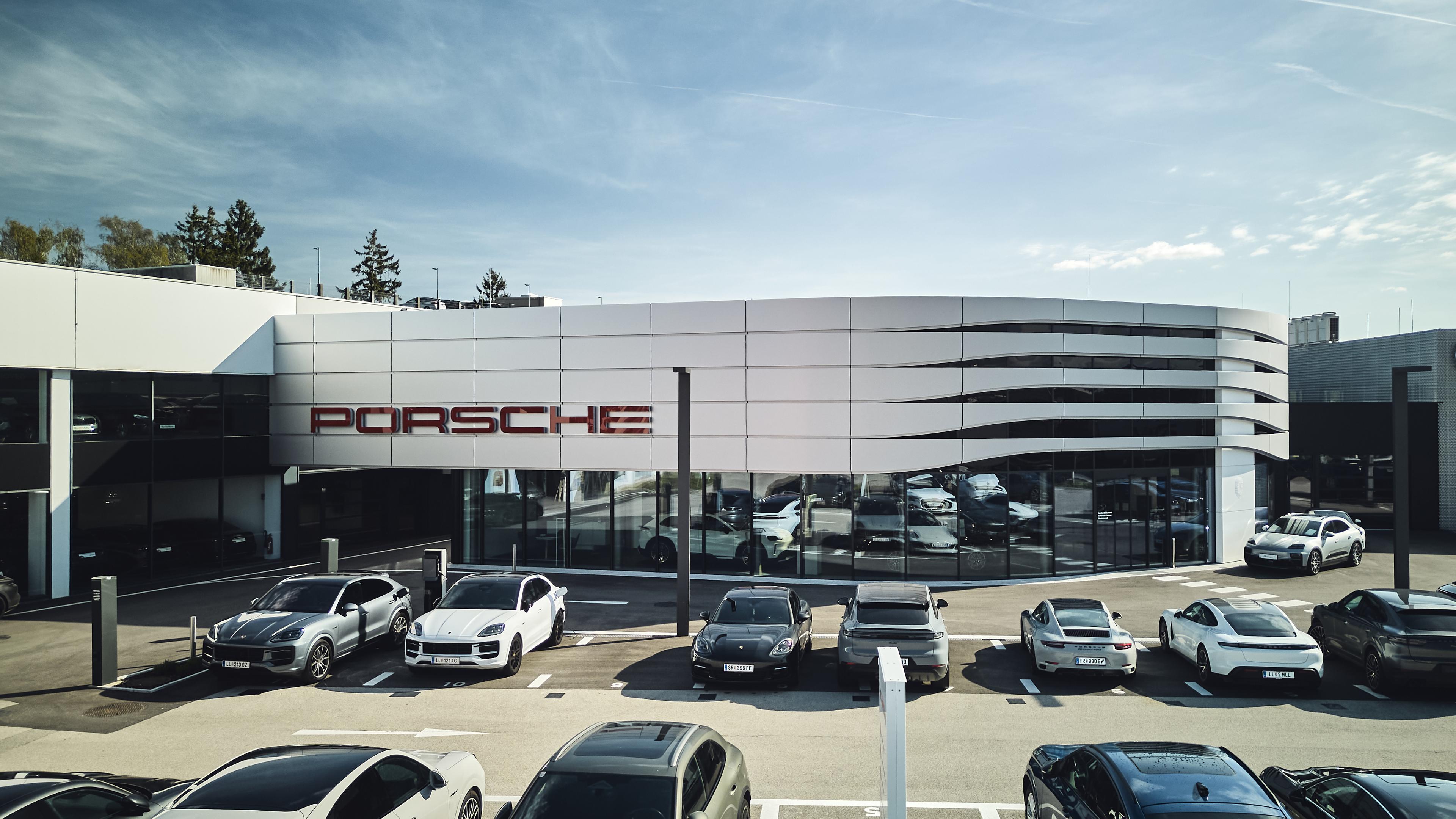 Modernes Porsche-Autohaus mit einem eleganten Außendesign, vor dem mehrere Porsche-Fahrzeuge unter einem klaren blauen Himmel geparkt sind.