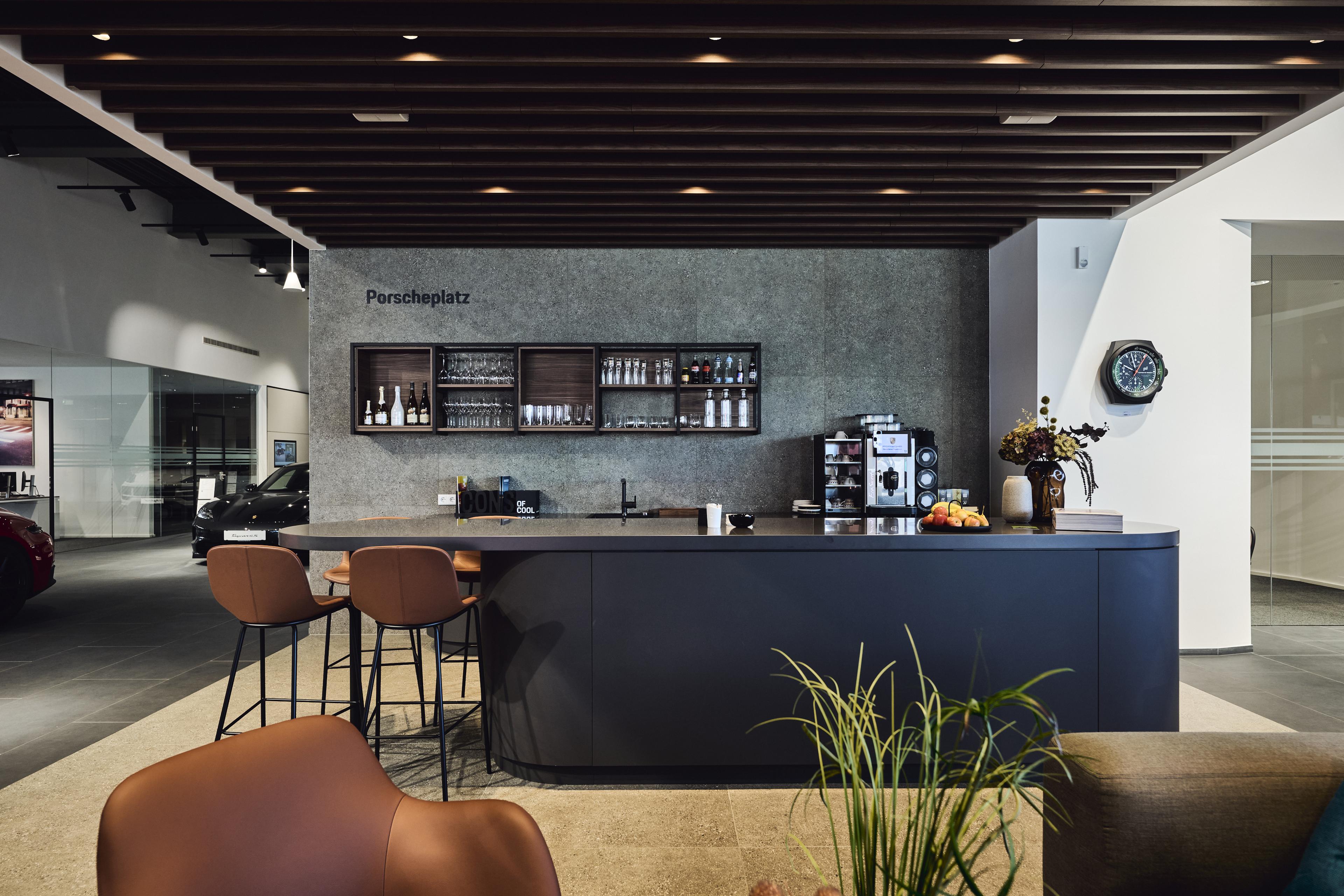 Eine moderne Cafébar mit Barhockern, Weingläsern, Flaschen, einer Kaffeemaschine, Obst und einem Blumenarrangement. Ein Schild trägt die Aufschrift „Porscheplatz.“
