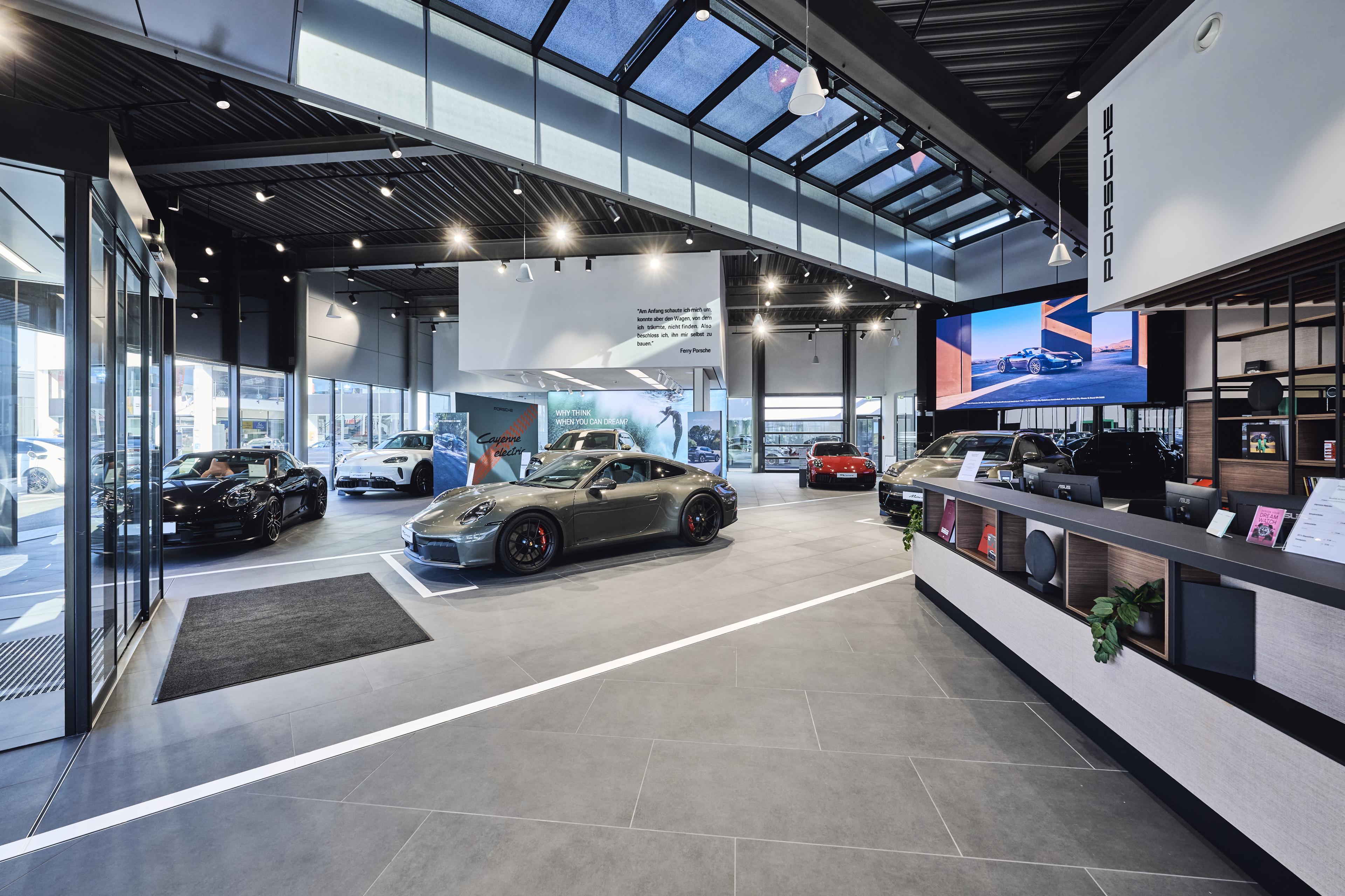 Ein moderner Auto-Showroom mit mehreren luxuriösen Sportwagen, darunter ein graues und ein rotes Modell, unter heller Deckenbeleuchtung und großen Fenstern.