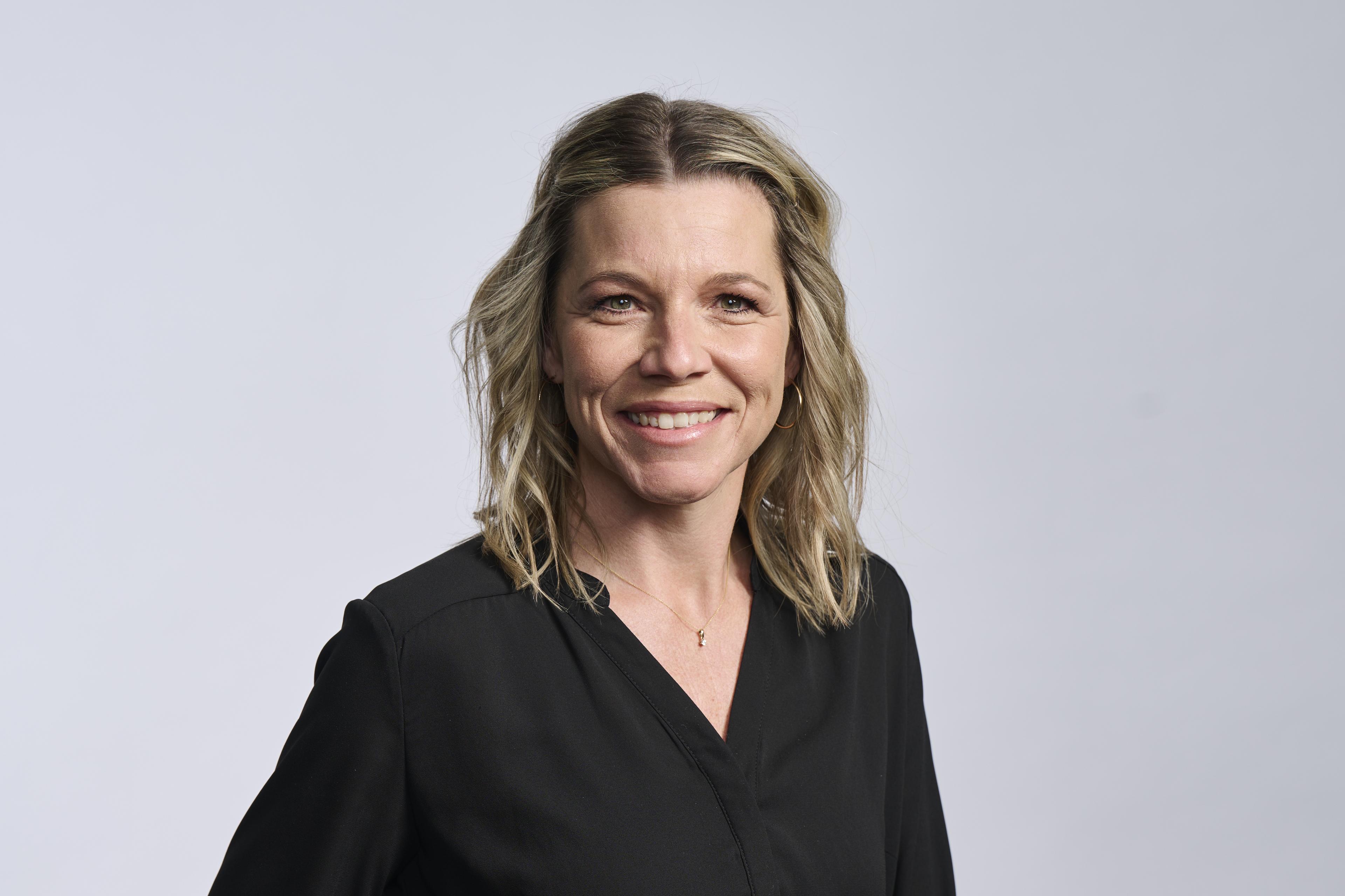 Lächelnde Frau mit schulterlangem blondem Haar, die eine schwarze Bluse und goldene Creolen trägt und vor einem schlichten Hintergrund steht.