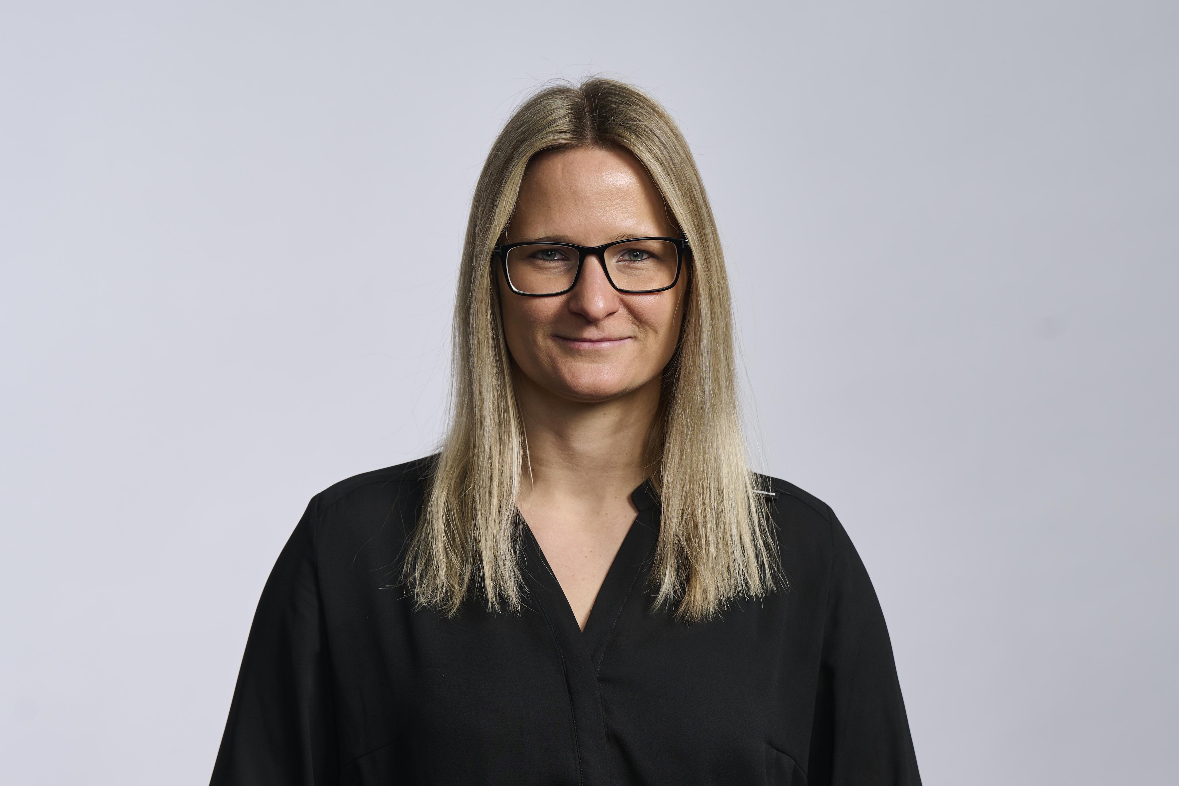 Eine Frau mit langen blonden Haaren und einer Brille, die ein schwarzes Shirt trägt und vor einem hellgrauen Hintergrund leicht lächelt.