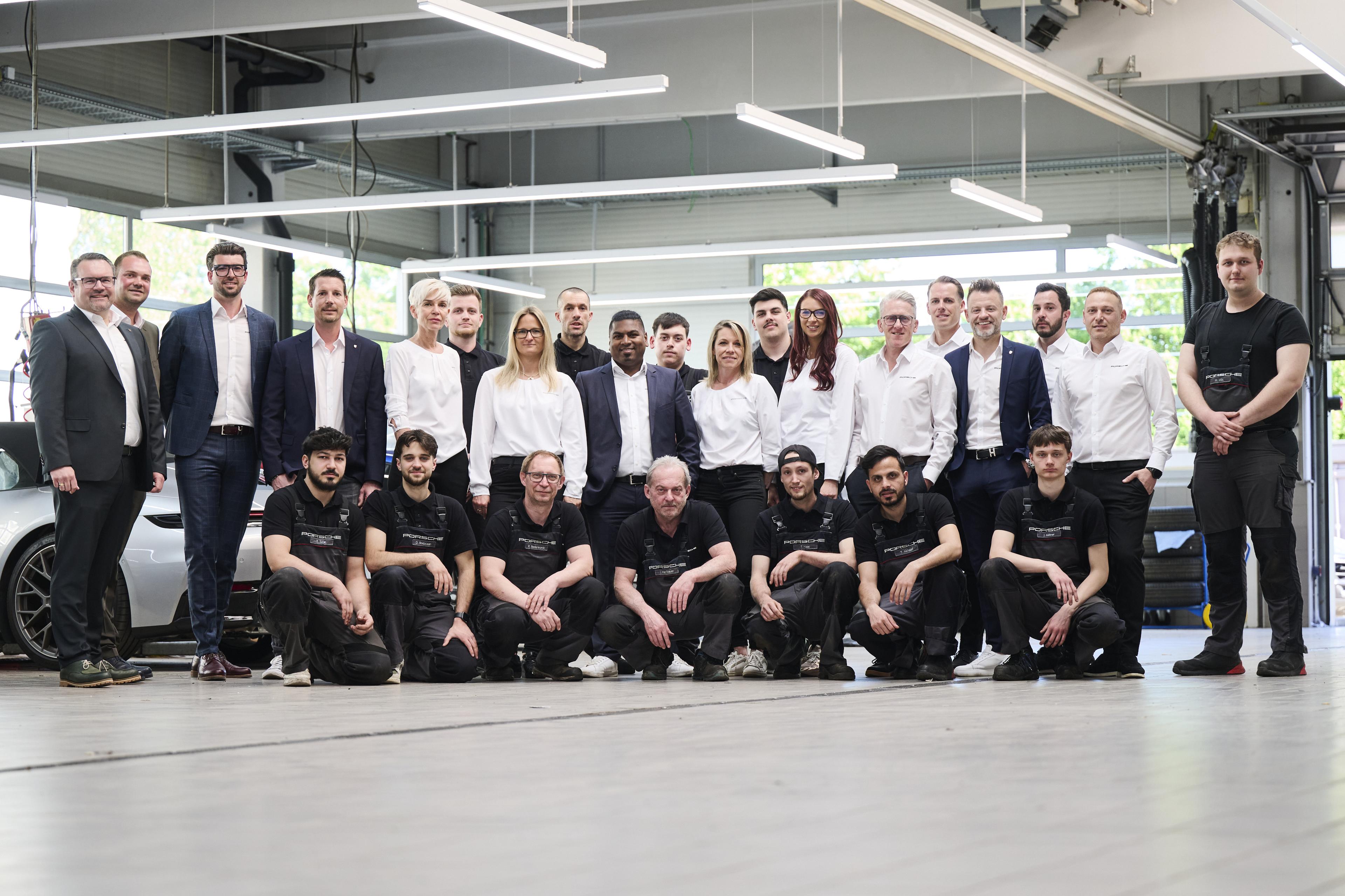 Ein Gruppenfoto von 22 Männern und Frauen in Business- und Arbeitskleidung, die in einer Kfz-Werkstatt mit Werkzeugen und Autos im Hintergrund für ein Foto posieren.