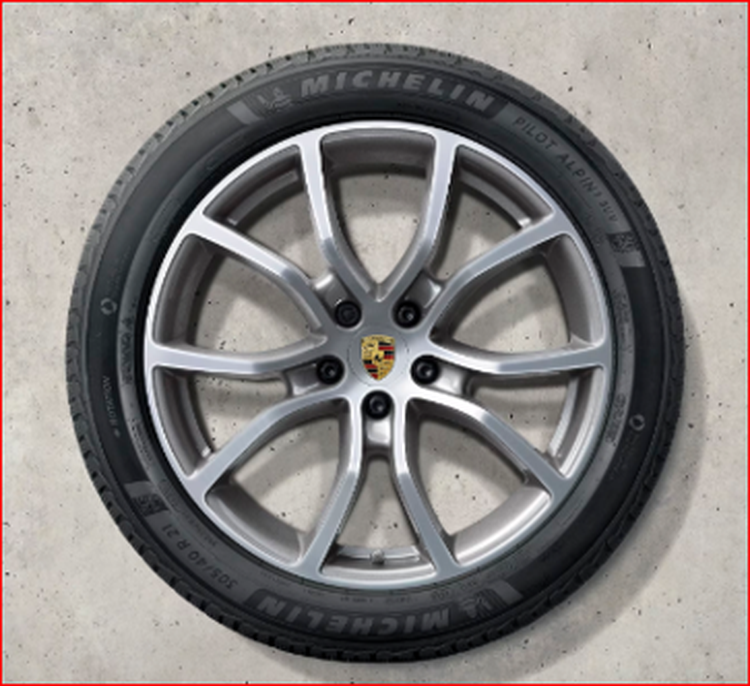 A Cayenne silver alloy wheel with a Porsche emblem at the center, mounted with a Michelin tire, on a concrete background.