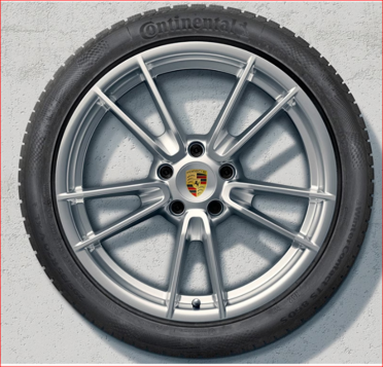 Porsche 911 Silver alloy wheel with a Porsche emblem in the center, encircled by a Continental tire, mounted against a gray wall.