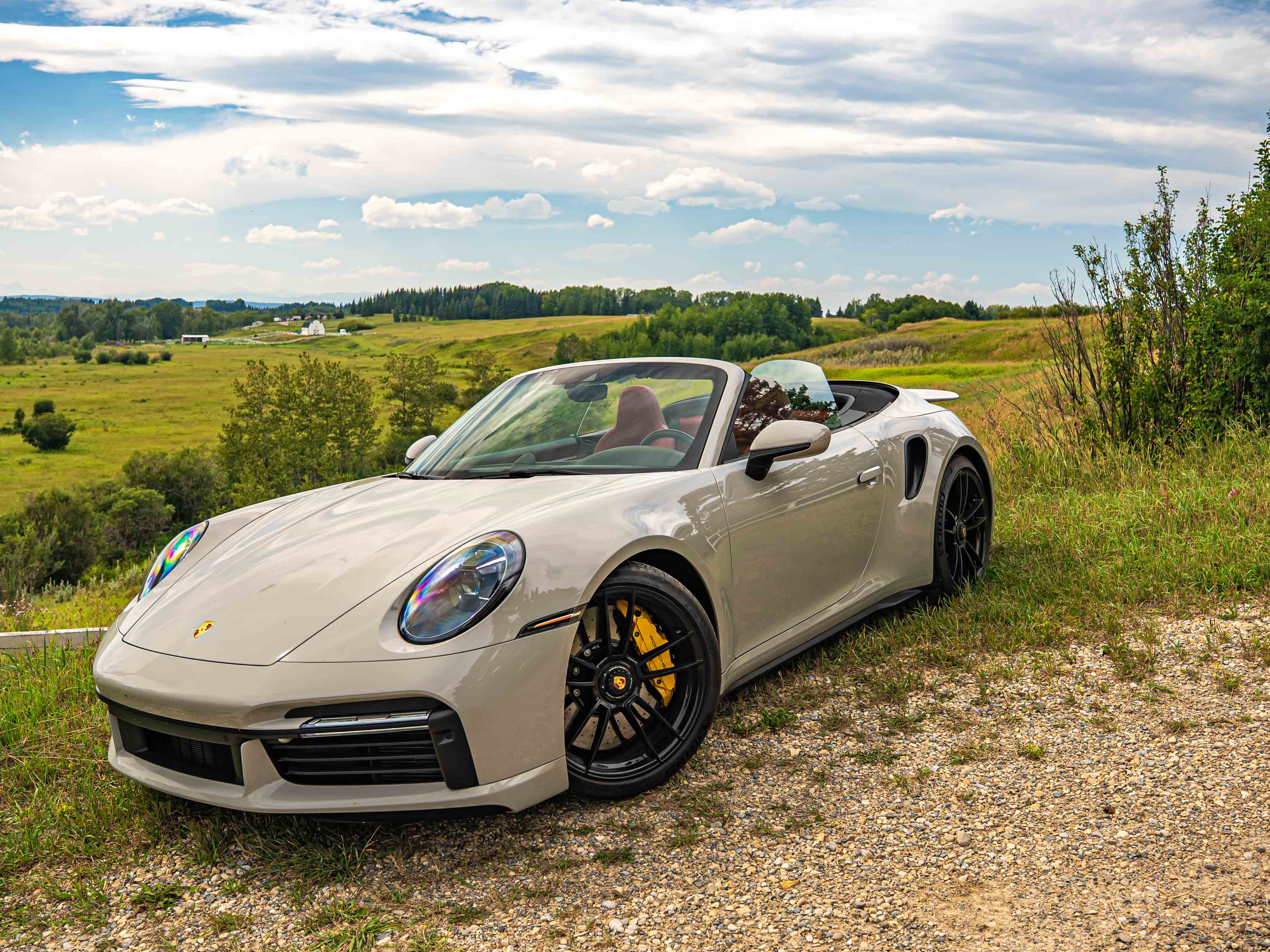 Chalk 911 Turbo S Cabriolet is parked on a gravel path, surrounded by lush green fields and trees under a partly cloudy sky.