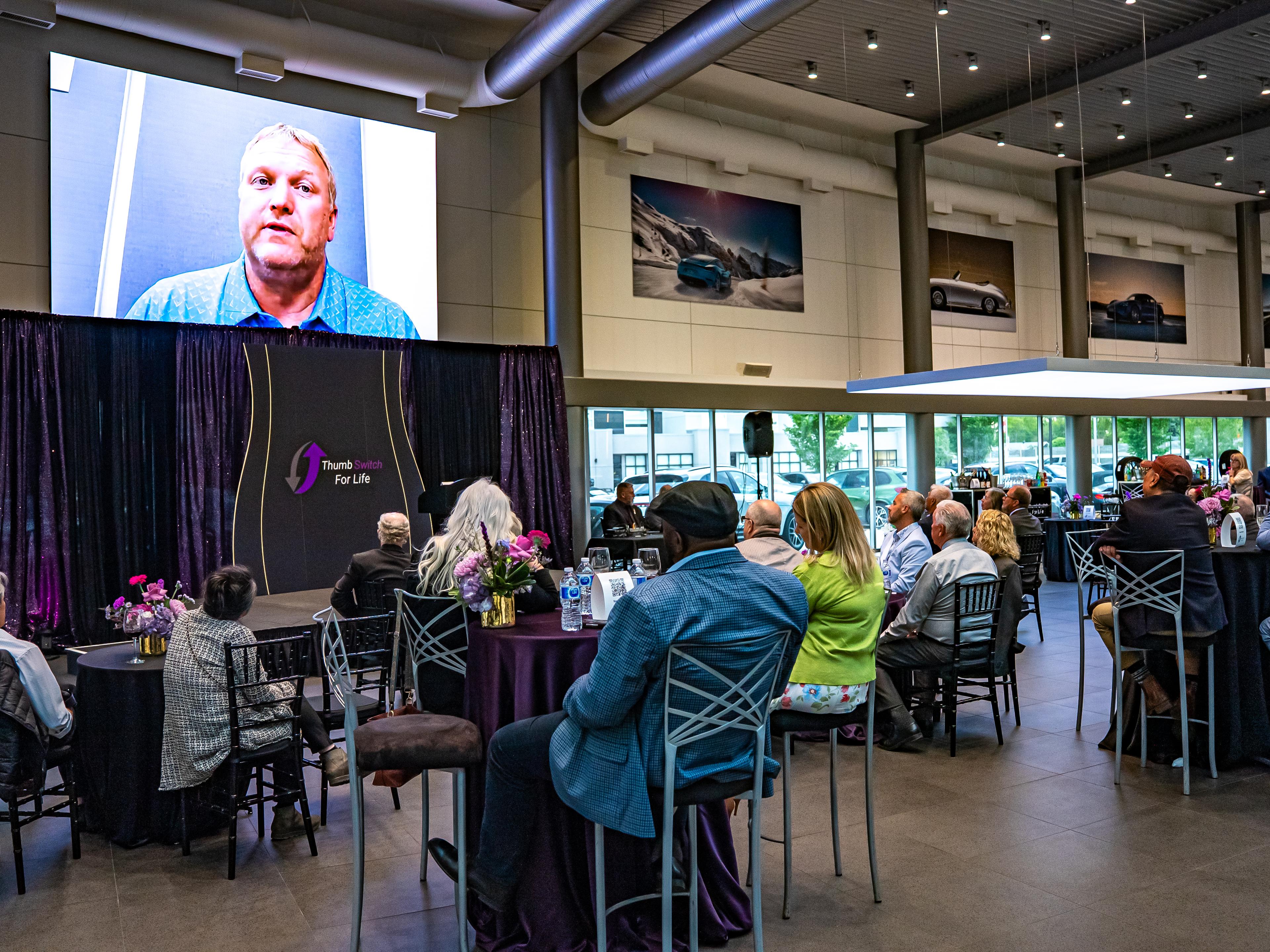A speaker on stage at an event titled "Thumb Switch For Life," with attendees seated at tables in a modern venue.