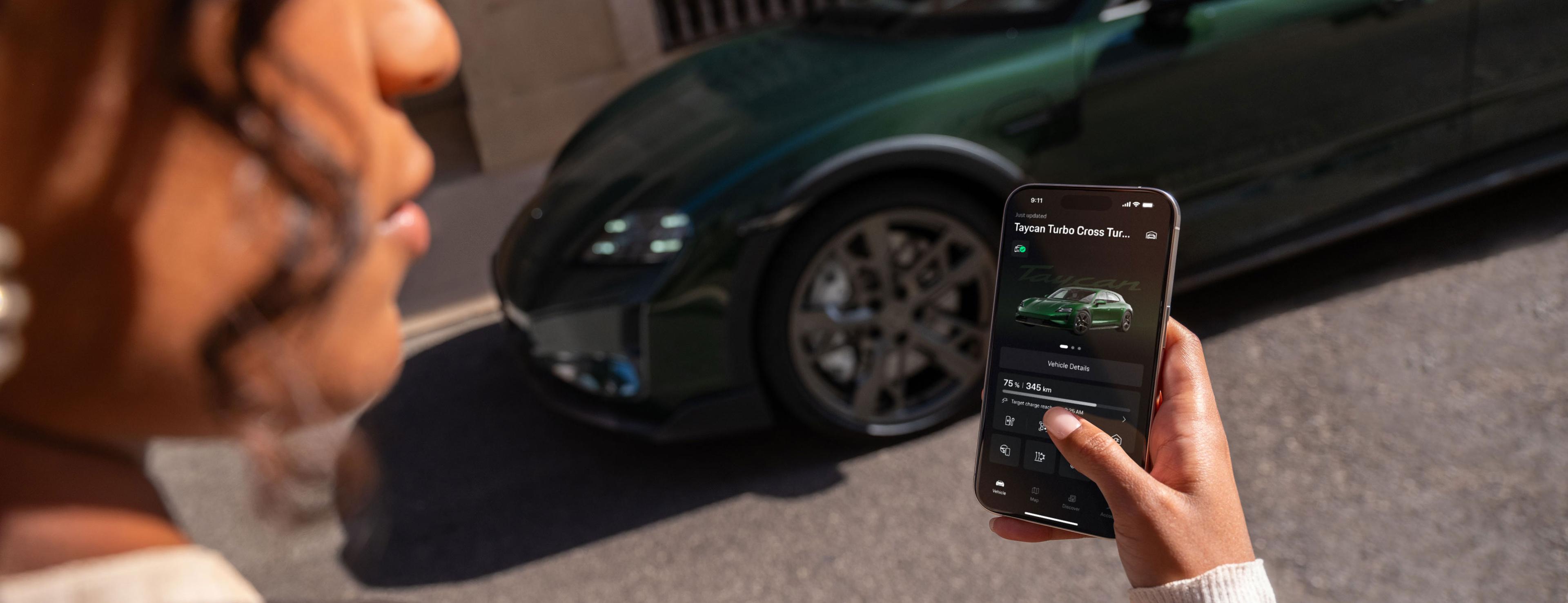 Eine Person steuert vor dem geparkten Fahrzeug ein grünes Auto mithilfe einer Smartphone-App, die Fahrzeugdetails und den Ladestand anzeigt.
