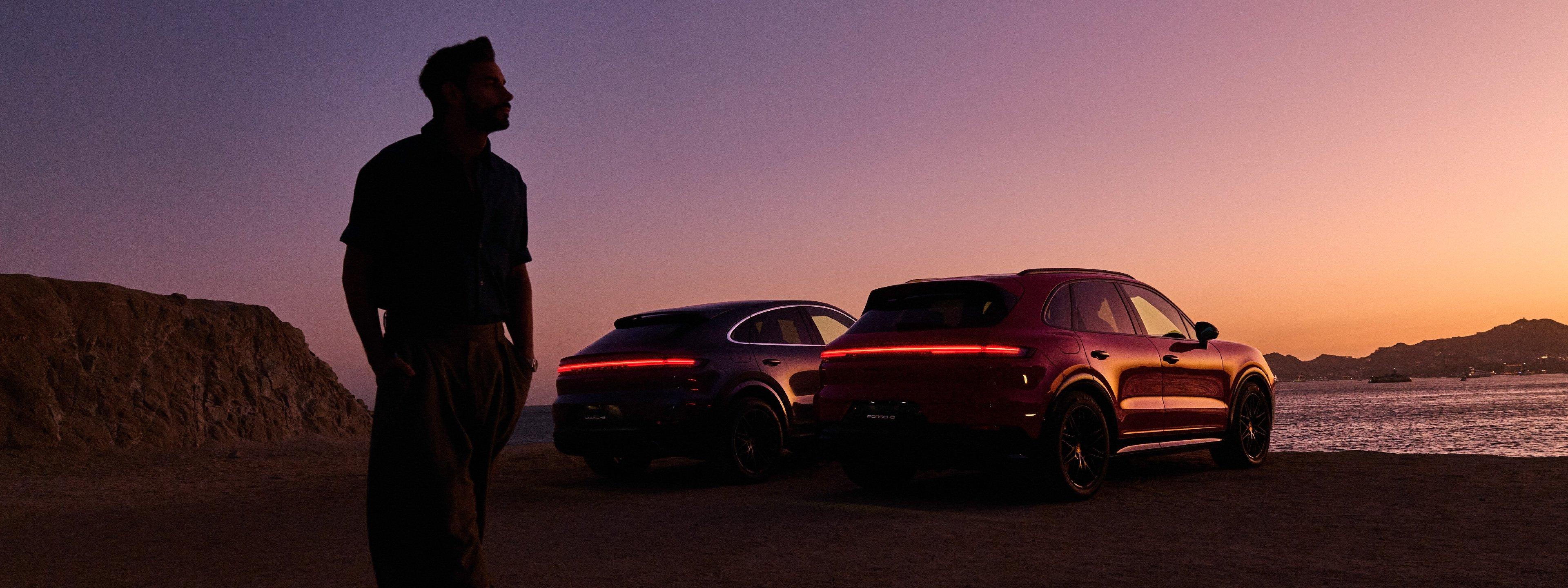 Silueta de un hombre viendo el atardecer en la playa con sus autos Porsche