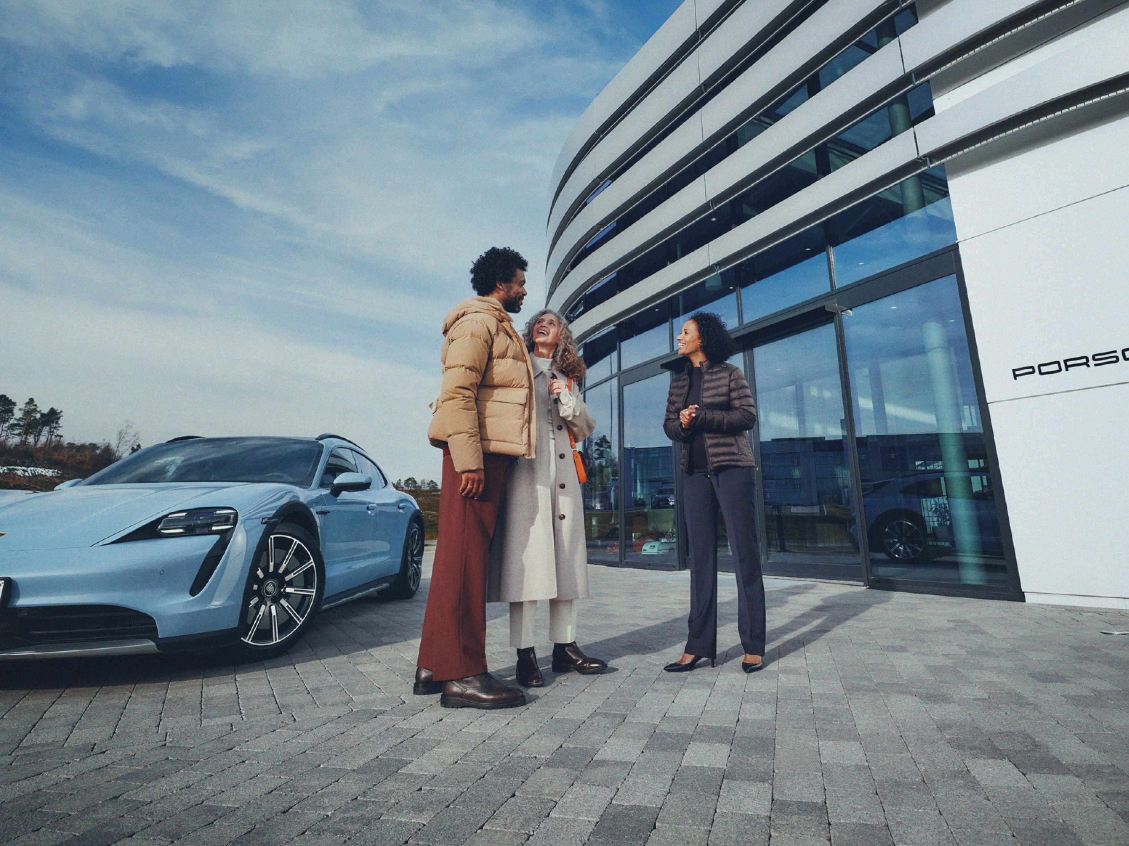 Tres personas conversando frente a un Porsche Taycan Cross Turismo azul y un concesionario Porsche moderno bajo un cielo azul.