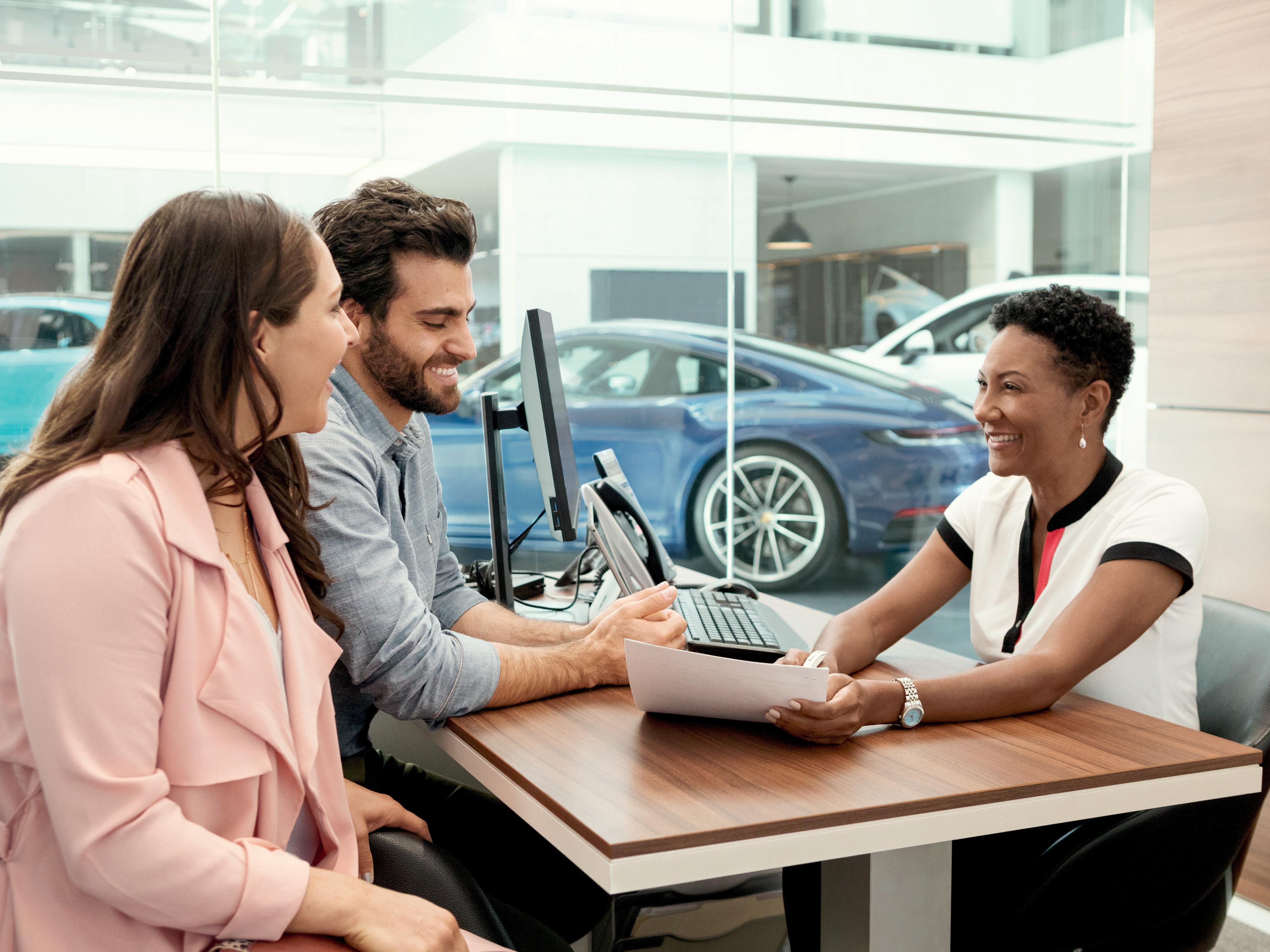 Una pareja sonriente interactúa con una vendedora en un concesionario de coches de lujo, con un Porsche azul deportivo visible en el fondo.