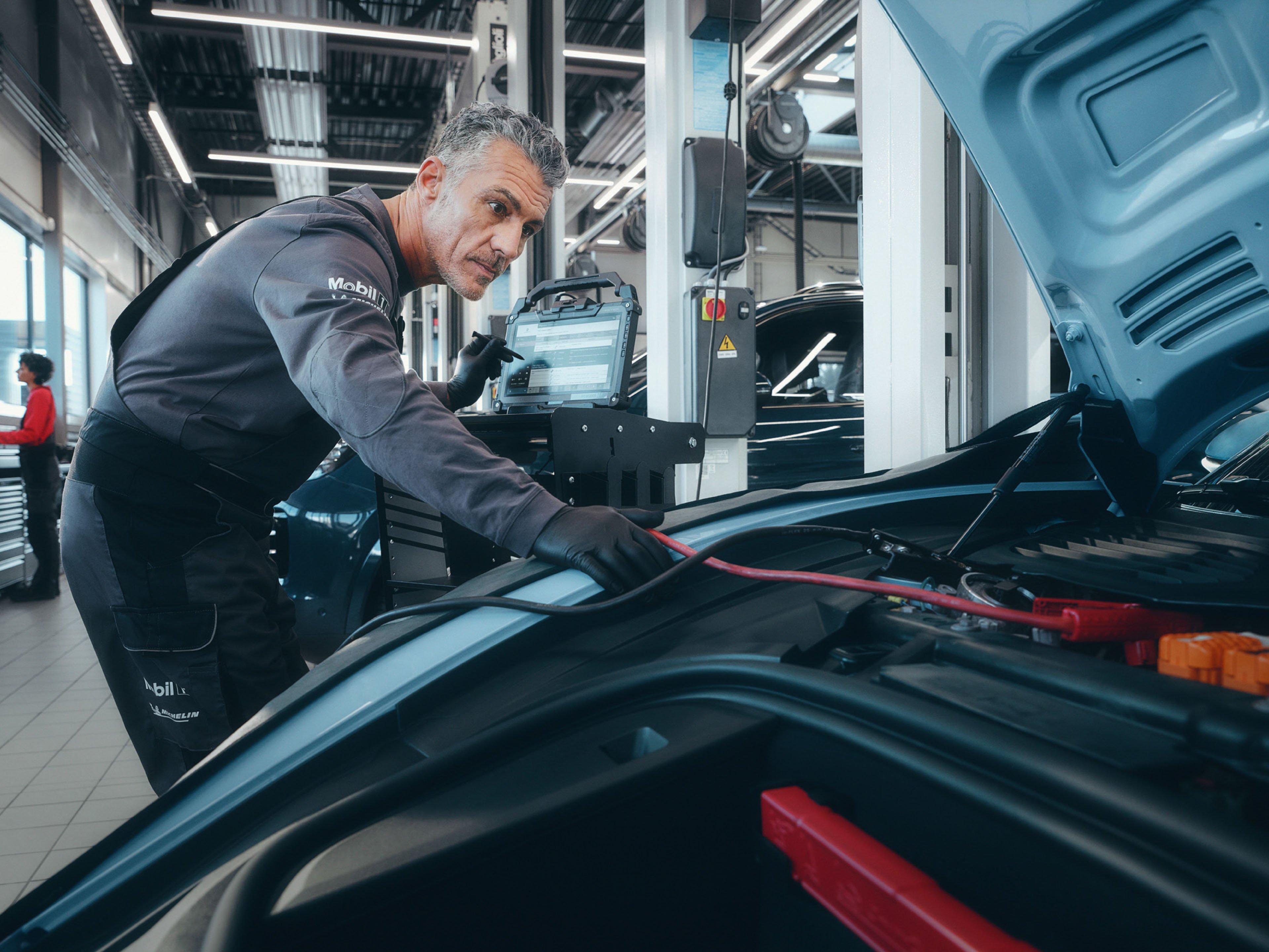 Un mecánico revisando el motor de un Porsche, con herramientas de diagnóstico y baterías de carga en un centro de servicio.