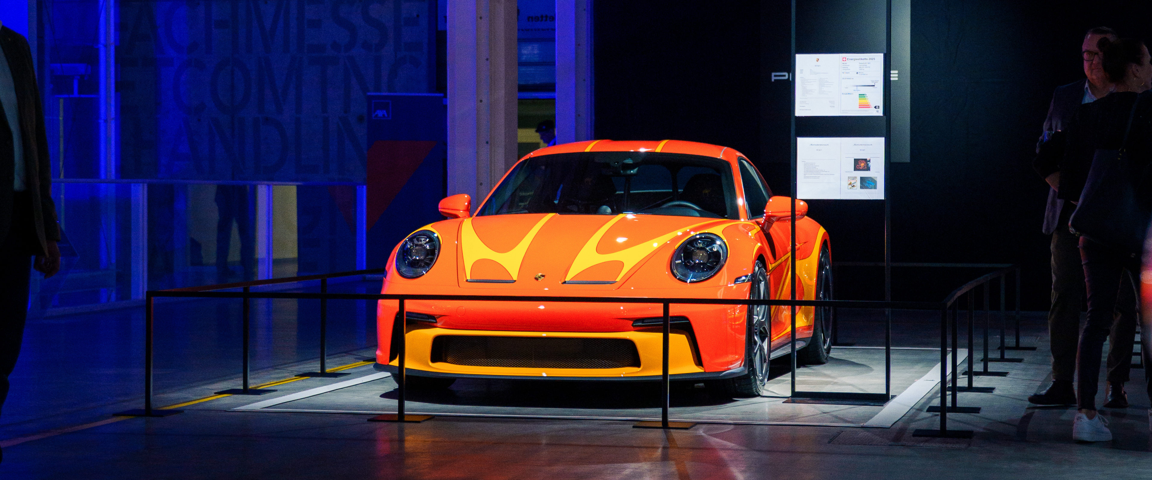 An orange sports car with flame-like yellow accents is displayed under spotlights, surrounded by a barrier in a dimly lit exhibition space.