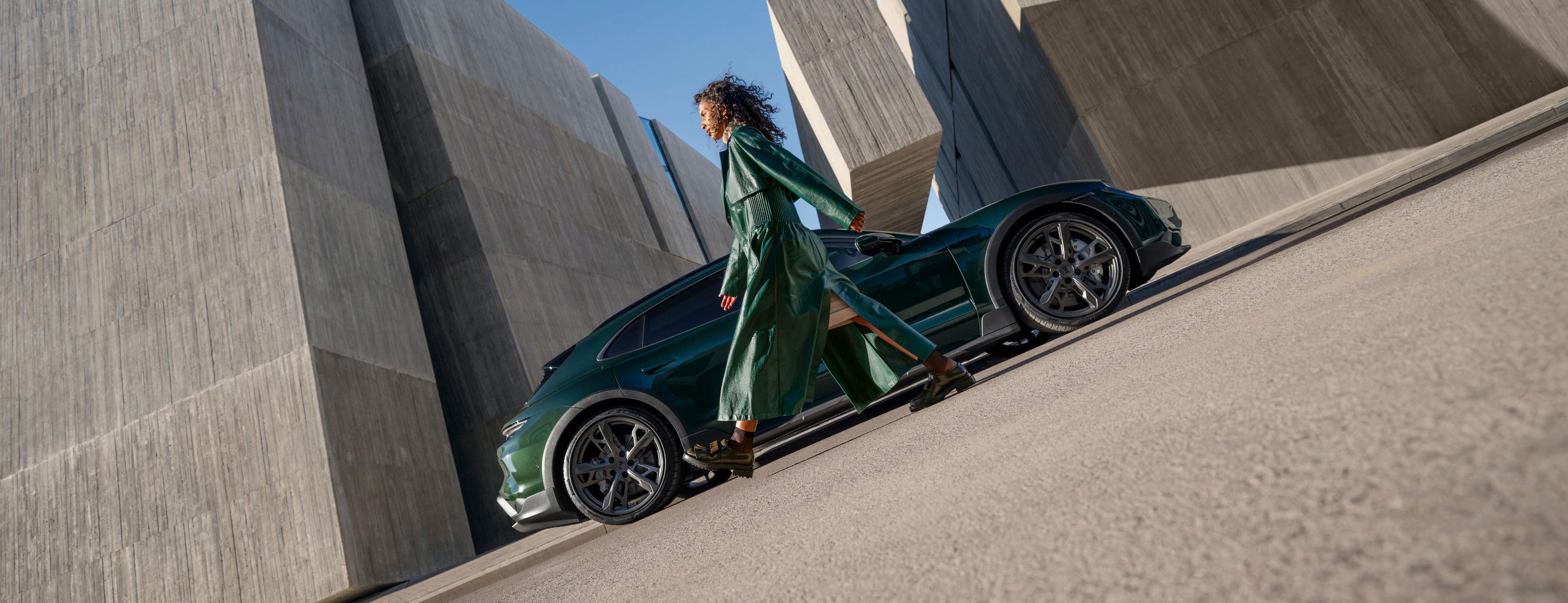 Eine Person in einem fließenden grünen Mantel geht an einem eleganten grünen Porsche Sportwagen vorbei.