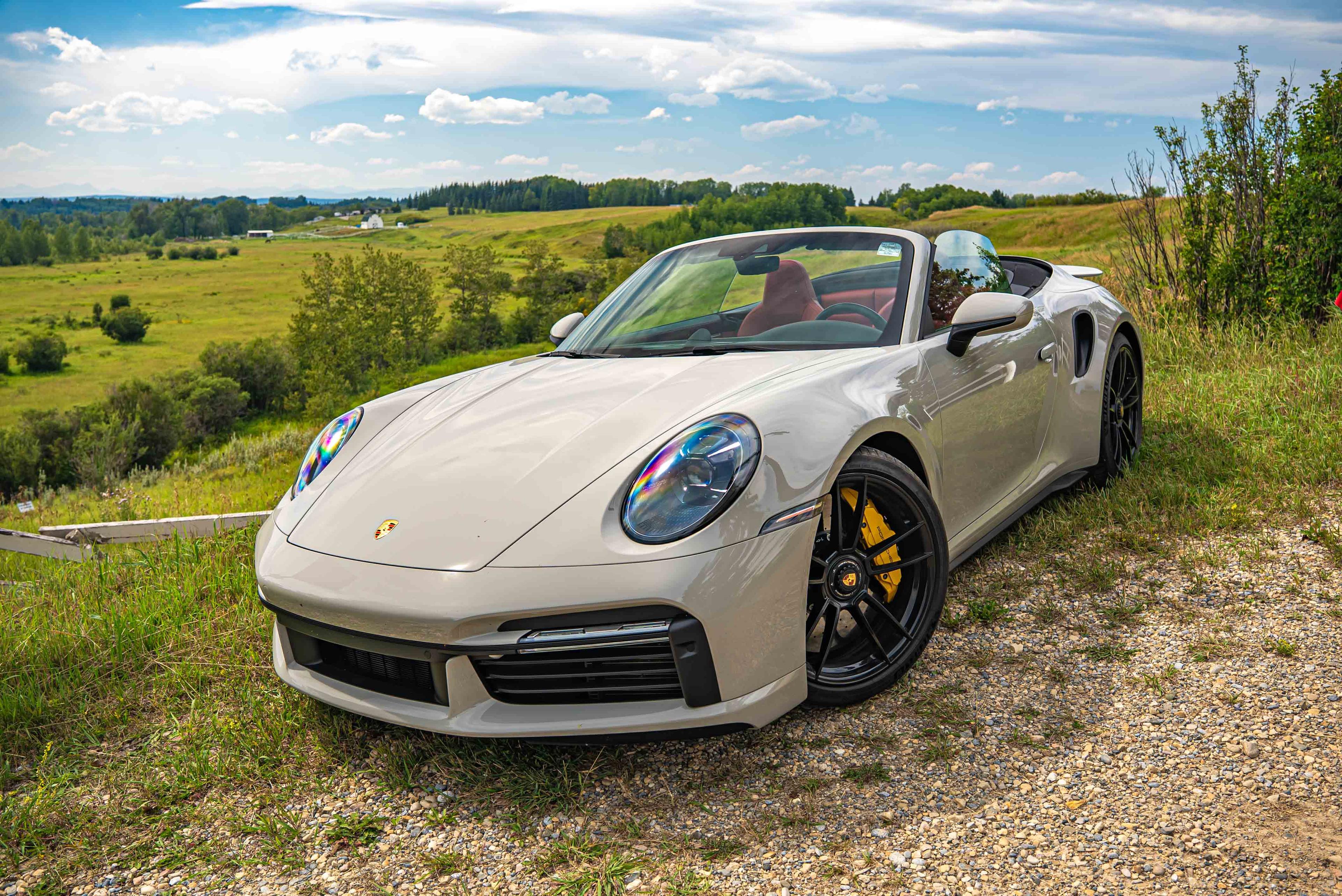 Chalk 911 Turbo S 992 Cabriolet is parked on a grassy hill with a scenic countryside view under a partly cloudy sky.