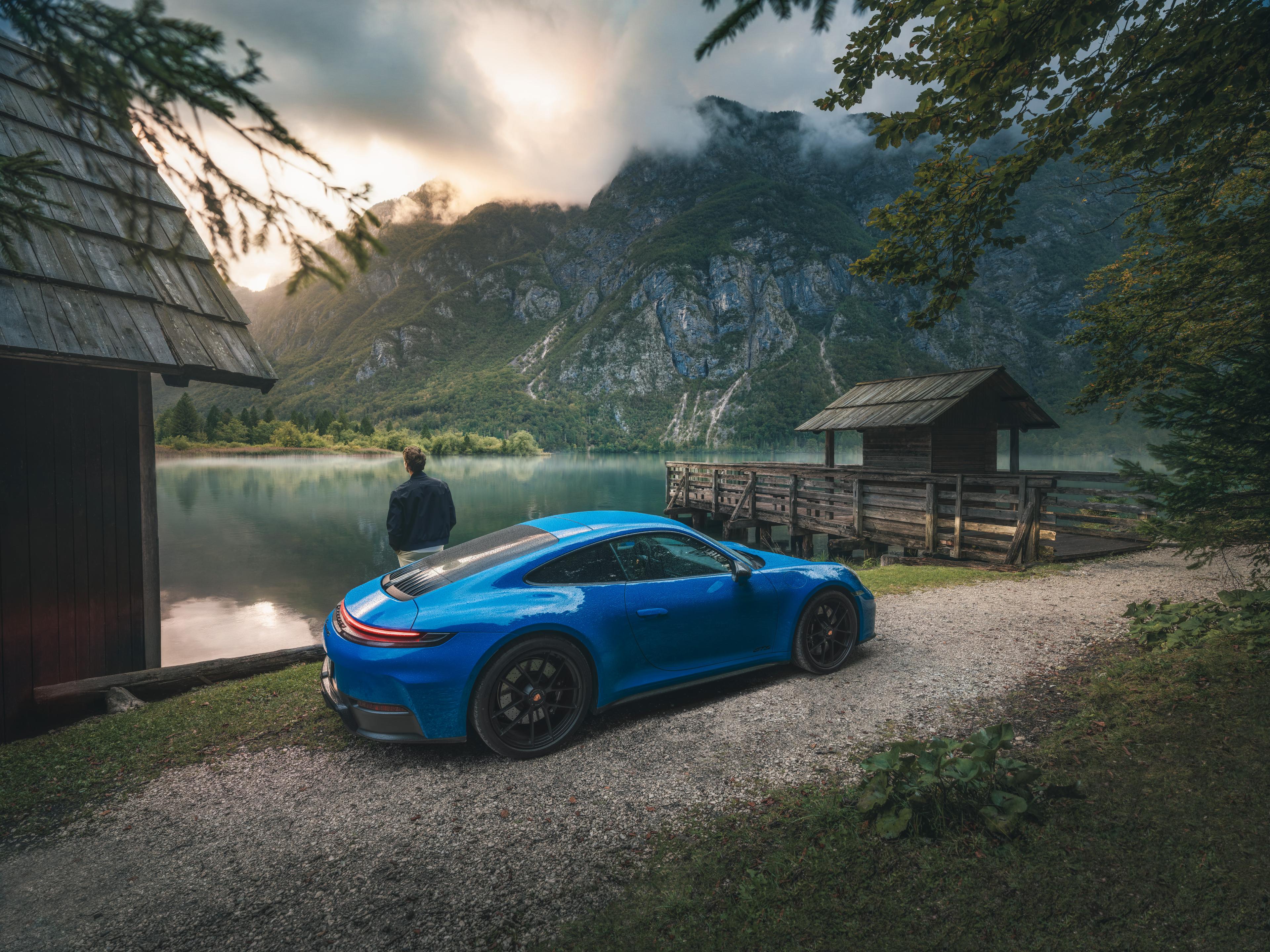 Ein blauer Porsche 911 ist in der Nähe eines ruhigen Sees geparkt, mit Bergen im Hintergrund. Eine Person steht bei Sonnenuntergang am Wasser neben einer Holzhütte.