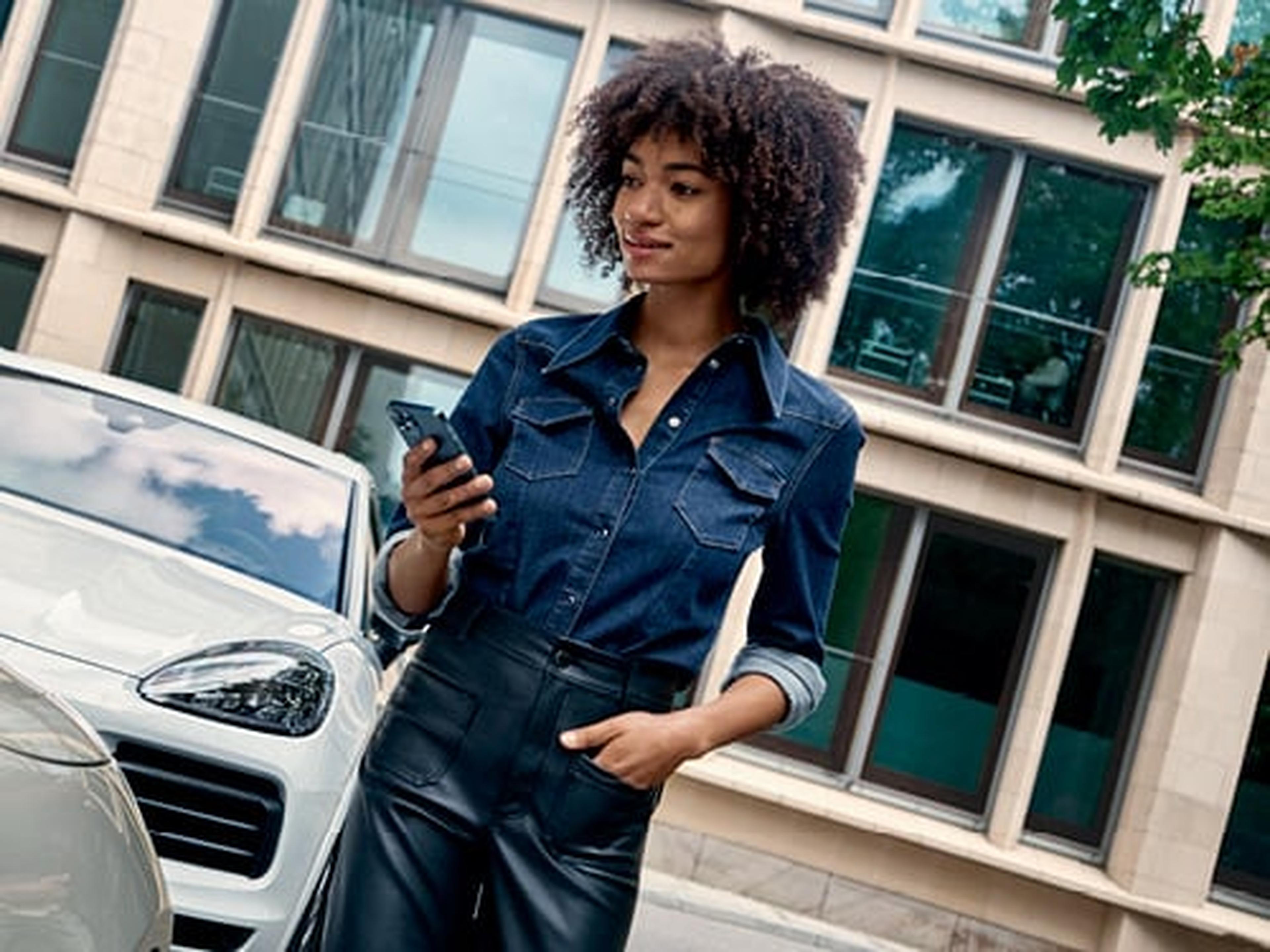 Joven mujer con camisa vaquera y pantalón de cuero sostiene un smartphone, con un Porsche Macan blanco parcialmente visible al fondo de un edificio moderno.