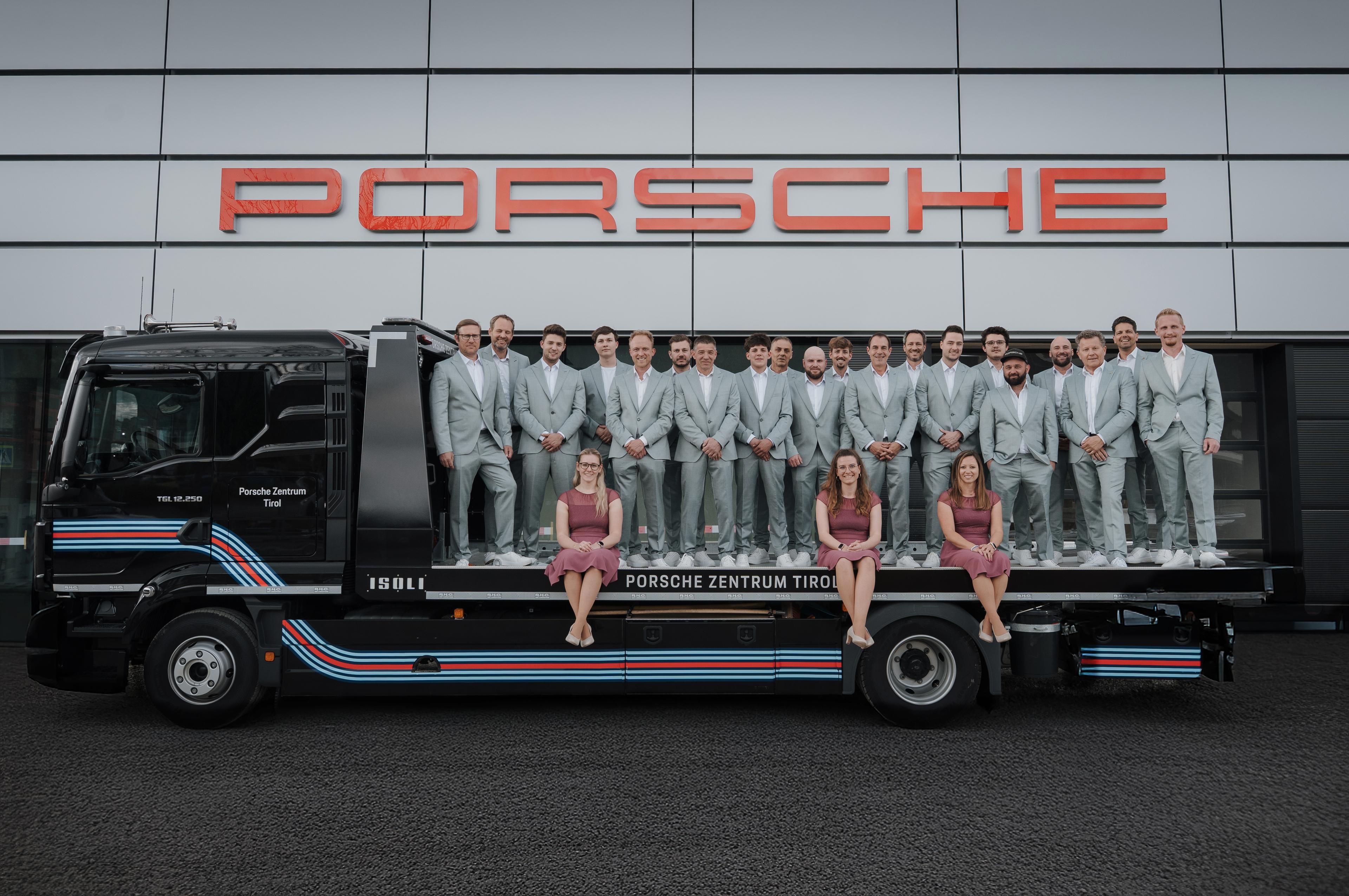 A group of people in matching gray suits and four in purple dresses pose on a truck bed in front of a building with a Porsche sign.