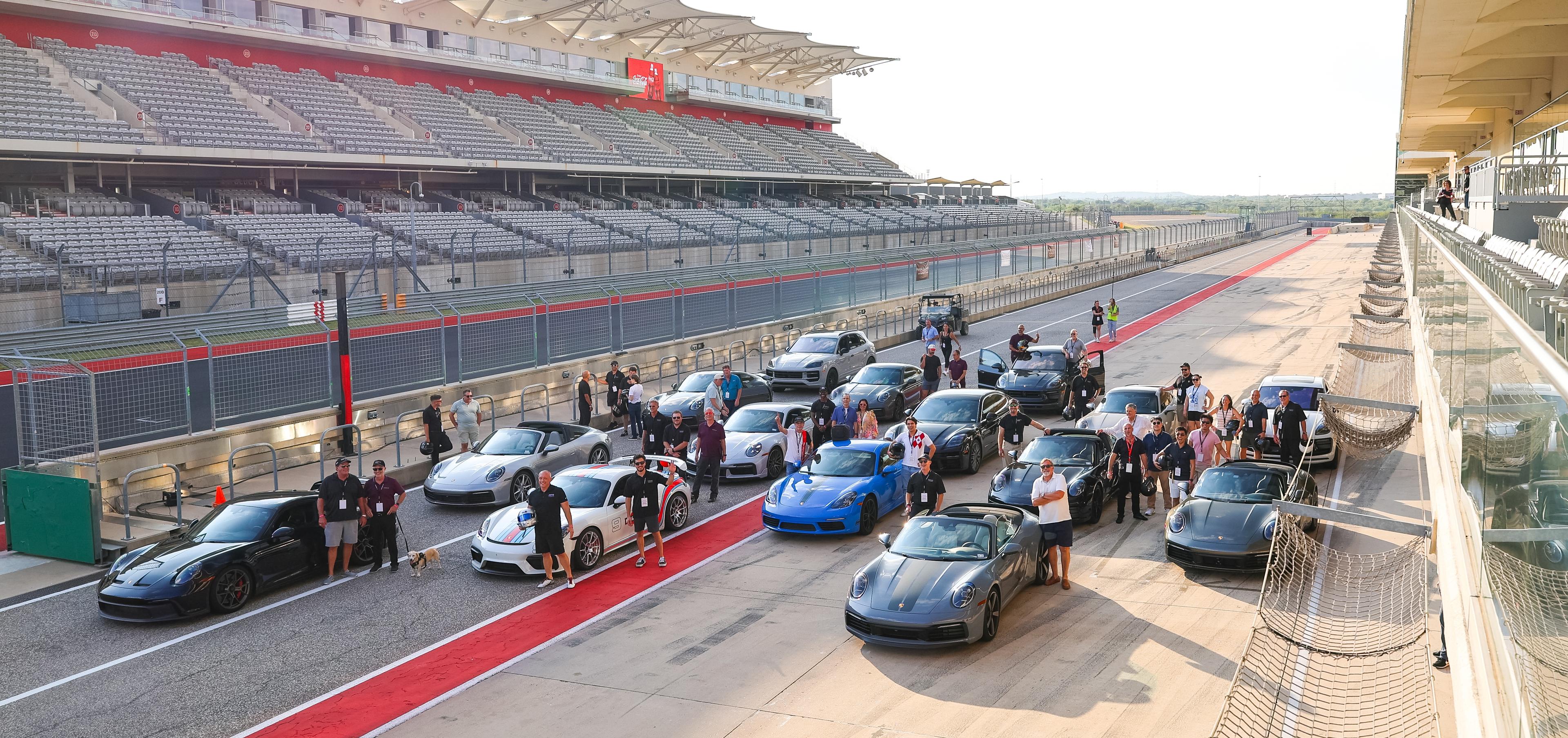 A group of people stands with several Porsche cars lined up on a racetrack pit lane, adjacent to an empty grandstand, under a clear sky.