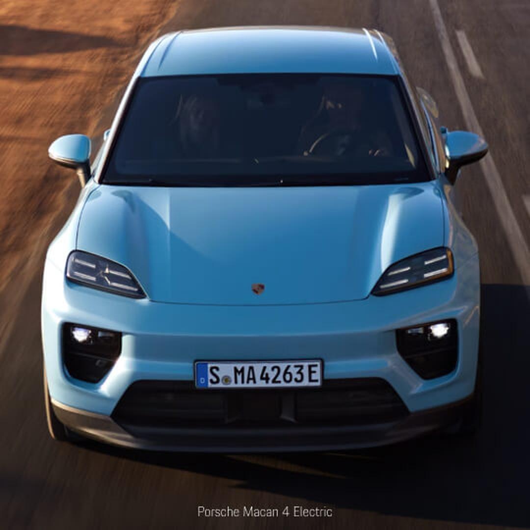High-angle front view of a Porsche Macan 4 Electric in Blue cruising on a sunny road.