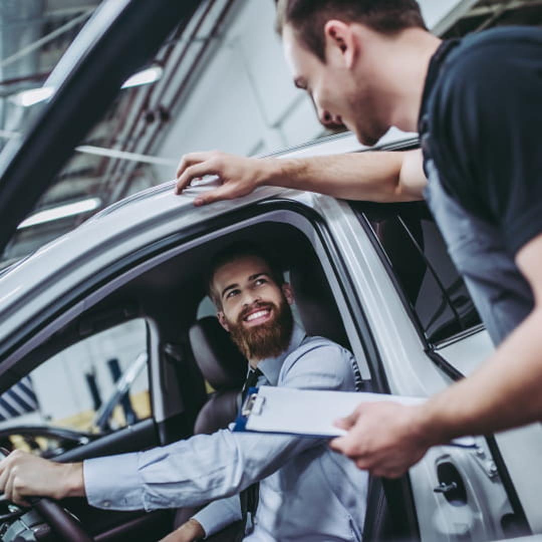 Automotive service technician speaking with a smiling customer seated inside a vehicle.