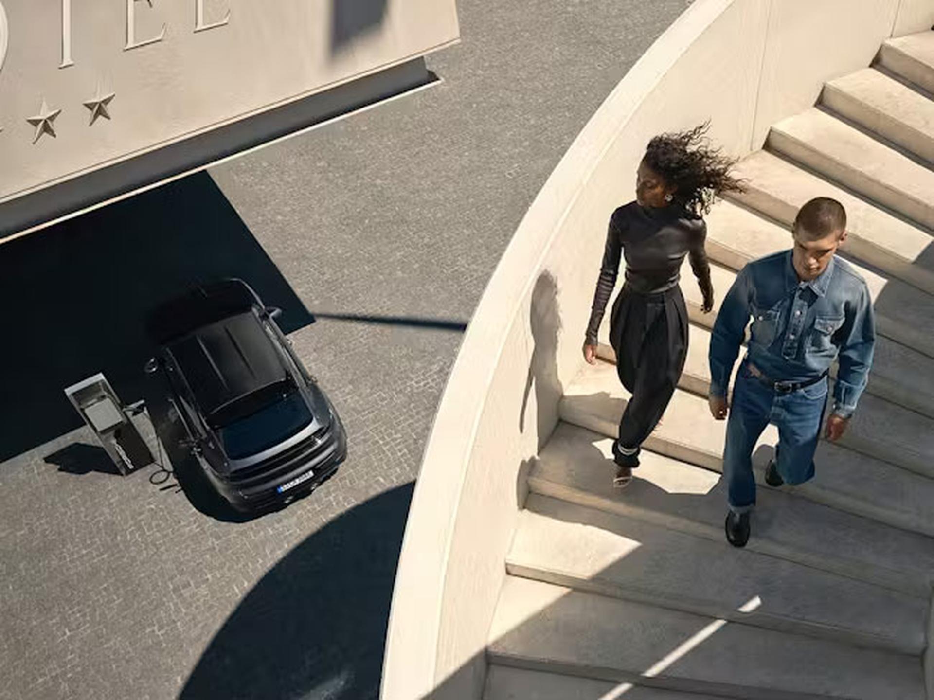 Ein Mann und eine Frau gehen eine geschwungene Treppe zu einem modernen Gebäude hinauf; weiter unten sind ein geparktes Auto und eine Ladestation zu sehen.