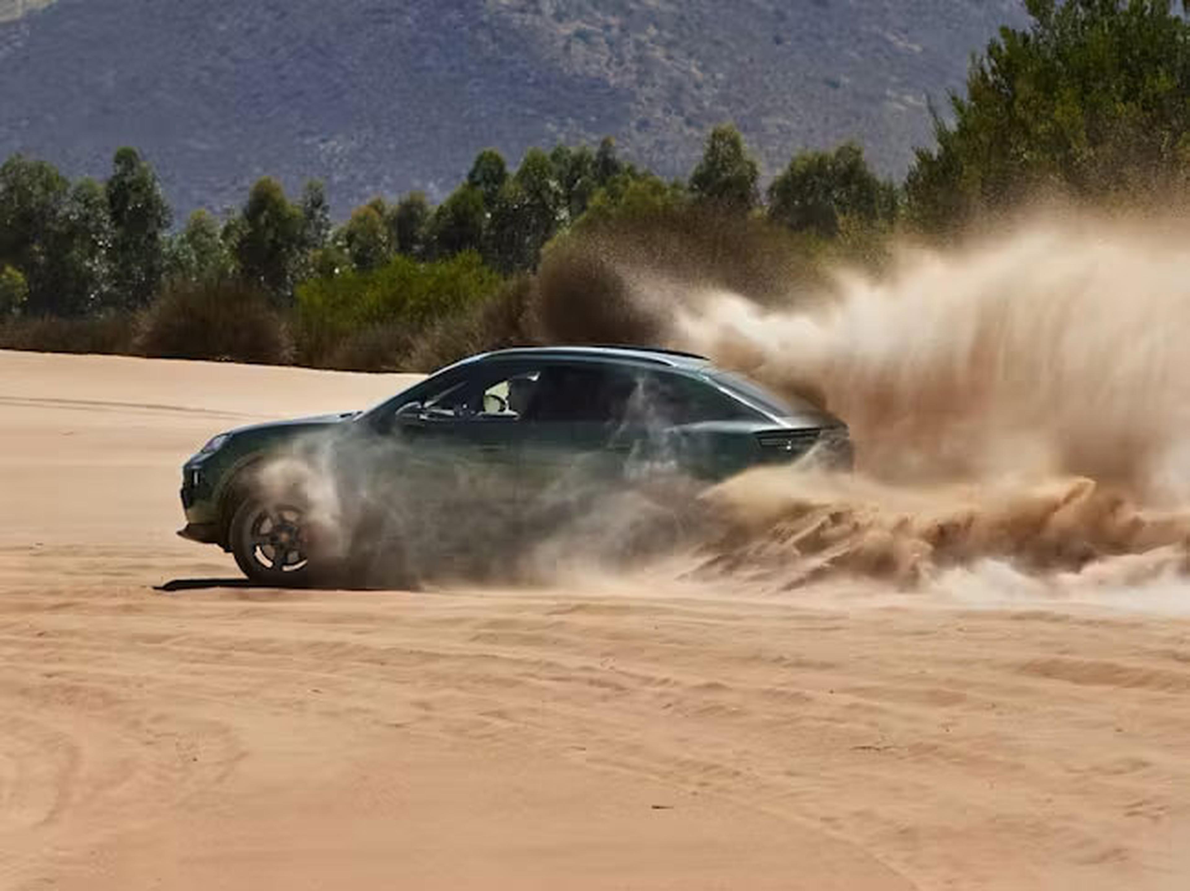Ein grüner SUV vollzieht eine scharfe Kurve in einer Sandwüste und wirbelt dabei eine große Staubwolke auf – vor der Kulisse von Bäumen und Bergen.