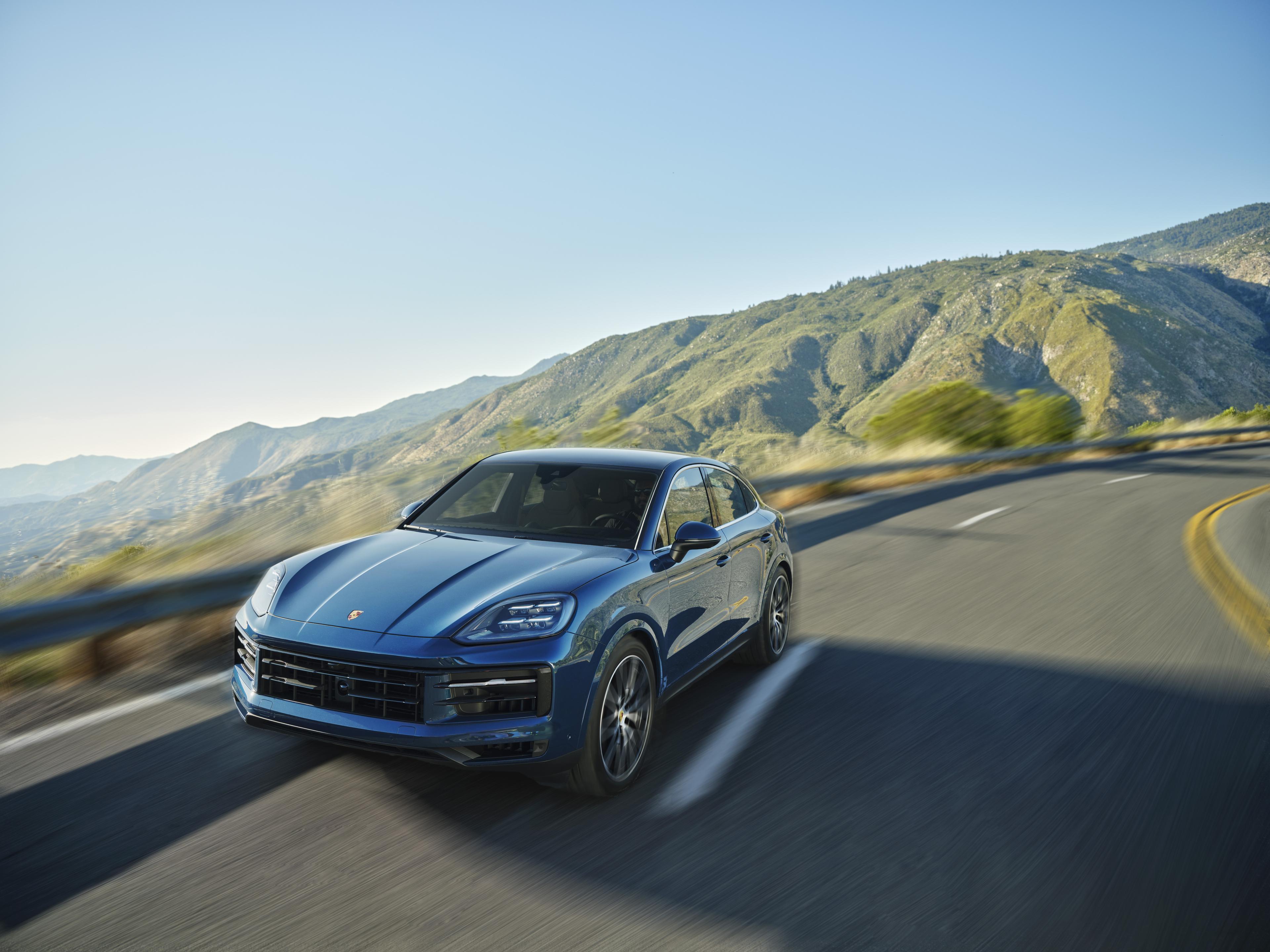 A blue 2023 Cayenne S Coupé drives on a scenic mountain road, with blurred motion and green hills in the background under a clear blue sky.