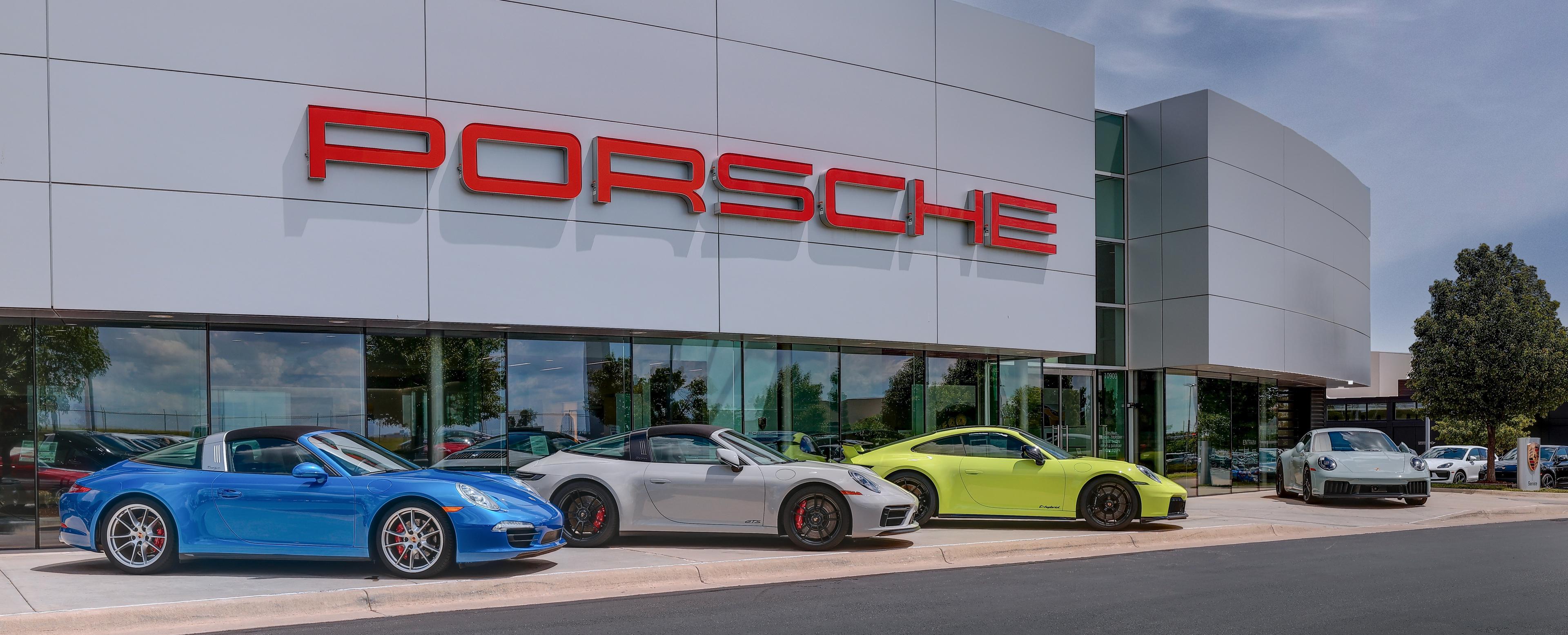 Four Porsche cars in blue, silver, and lime green are parked outside a Porsche dealership with a large sign on the building.