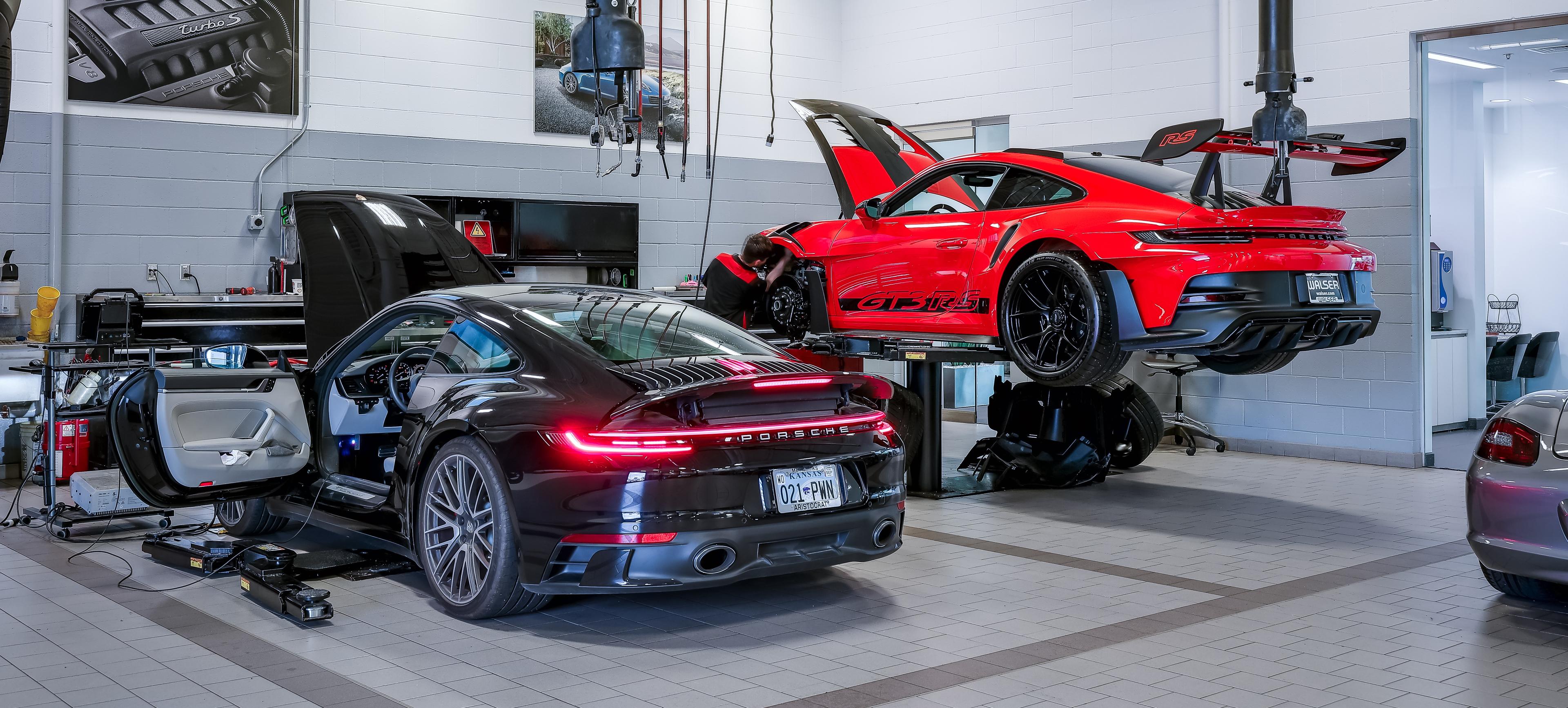 Two sports cars in an automotive workshop; a red car on a lift and a black car on the floor, both being serviced.