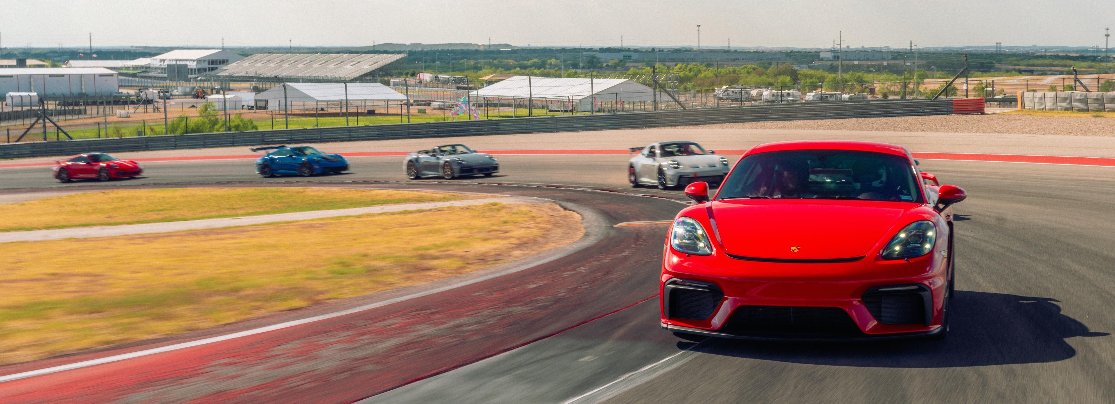 Red Porsche 718 Cayman sports car leading a group of vehicles around a race track, with grass and buildings in the background at Circuit of The Americas.