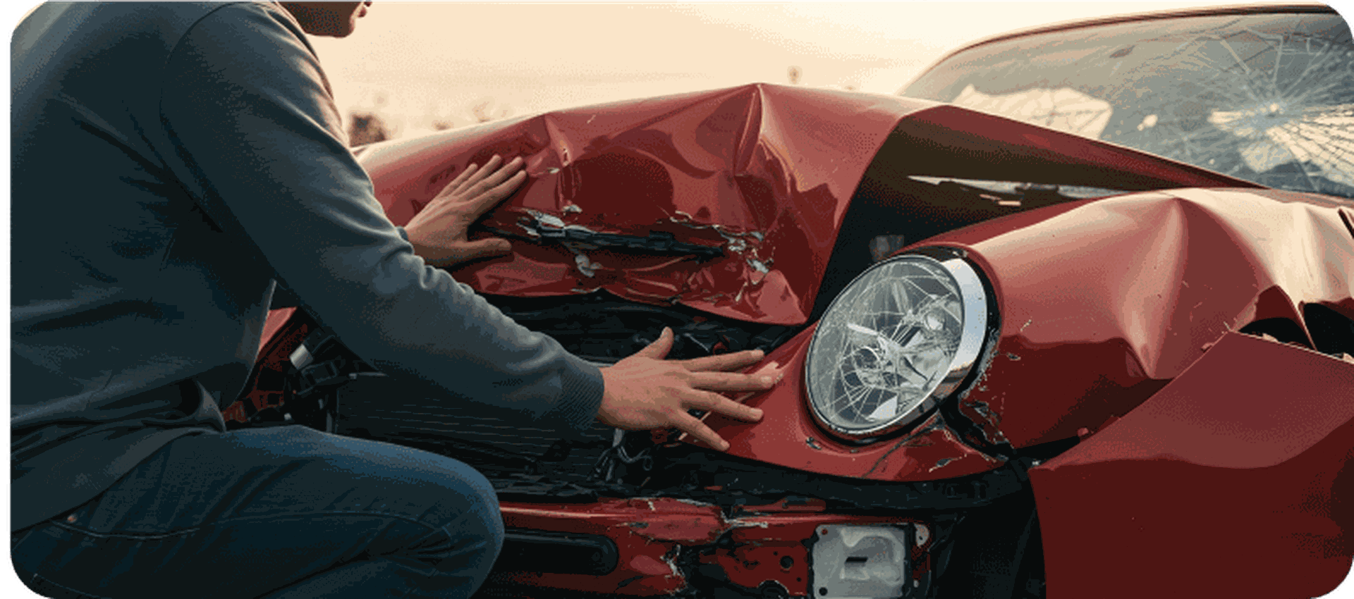 A person examines the heavily damaged front of a red car with a shattered headlight and crumpled hood.